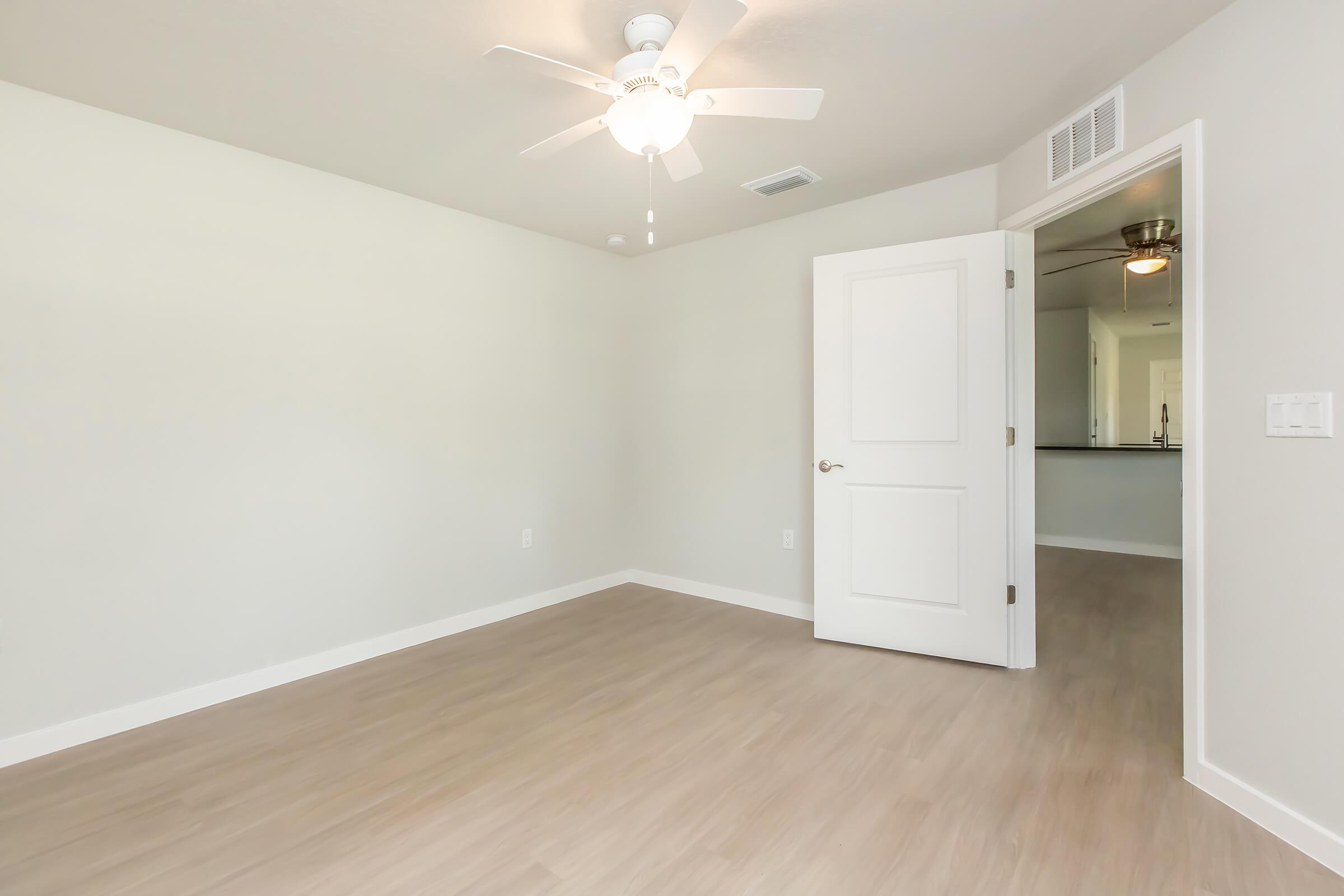 A brightly lit empty room with light gray walls and light wood flooring. A ceiling fan is mounted in the center, and a closed white door is visible on the right. In the background, an open door leads to another room with visible kitchen fixtures. The overall atmosphere is clean and modern.