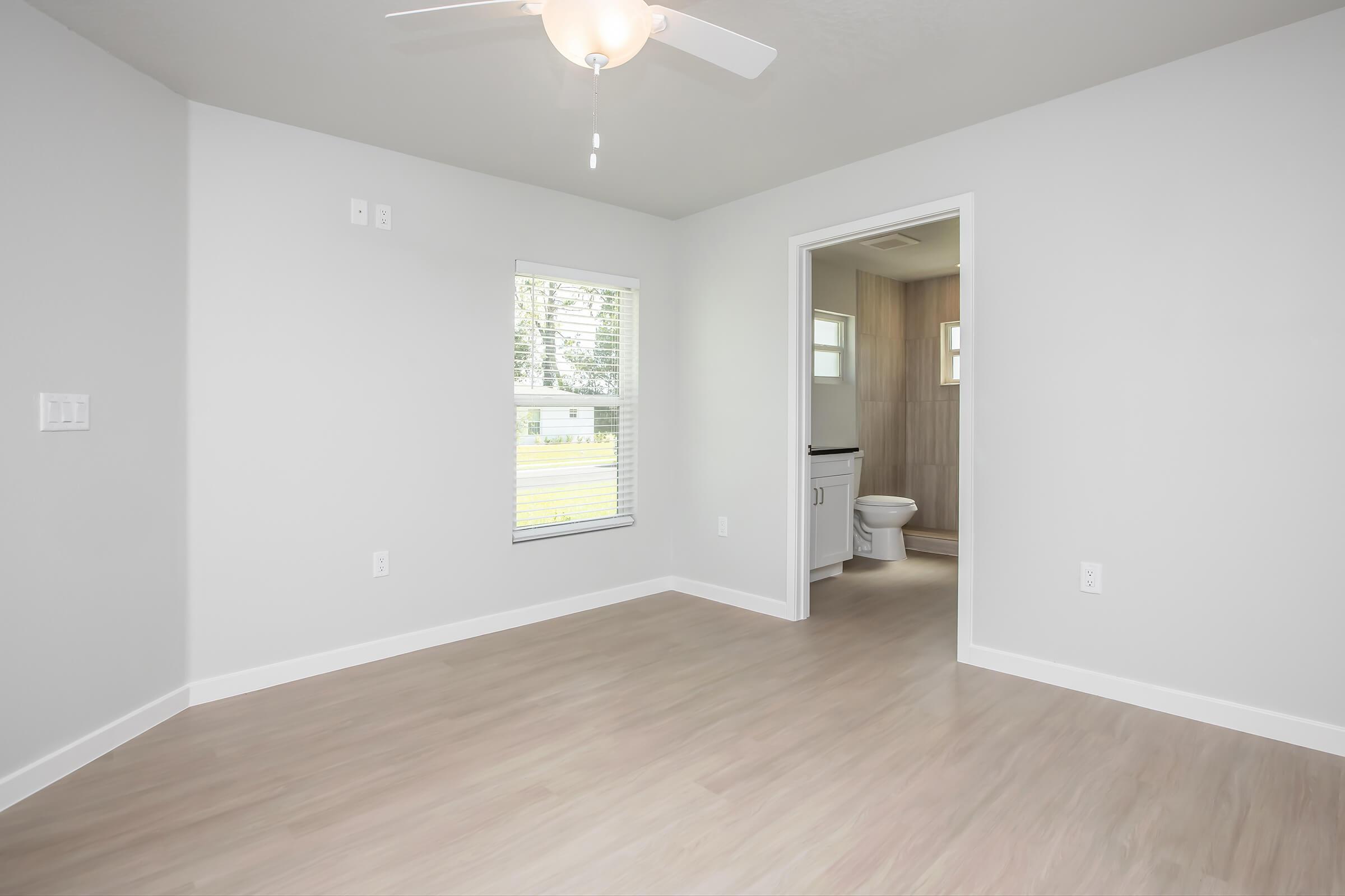 A clean, empty room with light gray walls and a ceiling fan. The room features a window allowing natural light, overlooking a green space outside. A doorway leads to a bathroom area with neutral-toned tiles. The flooring is light-colored, enhancing the spacious and airy feel of the room.