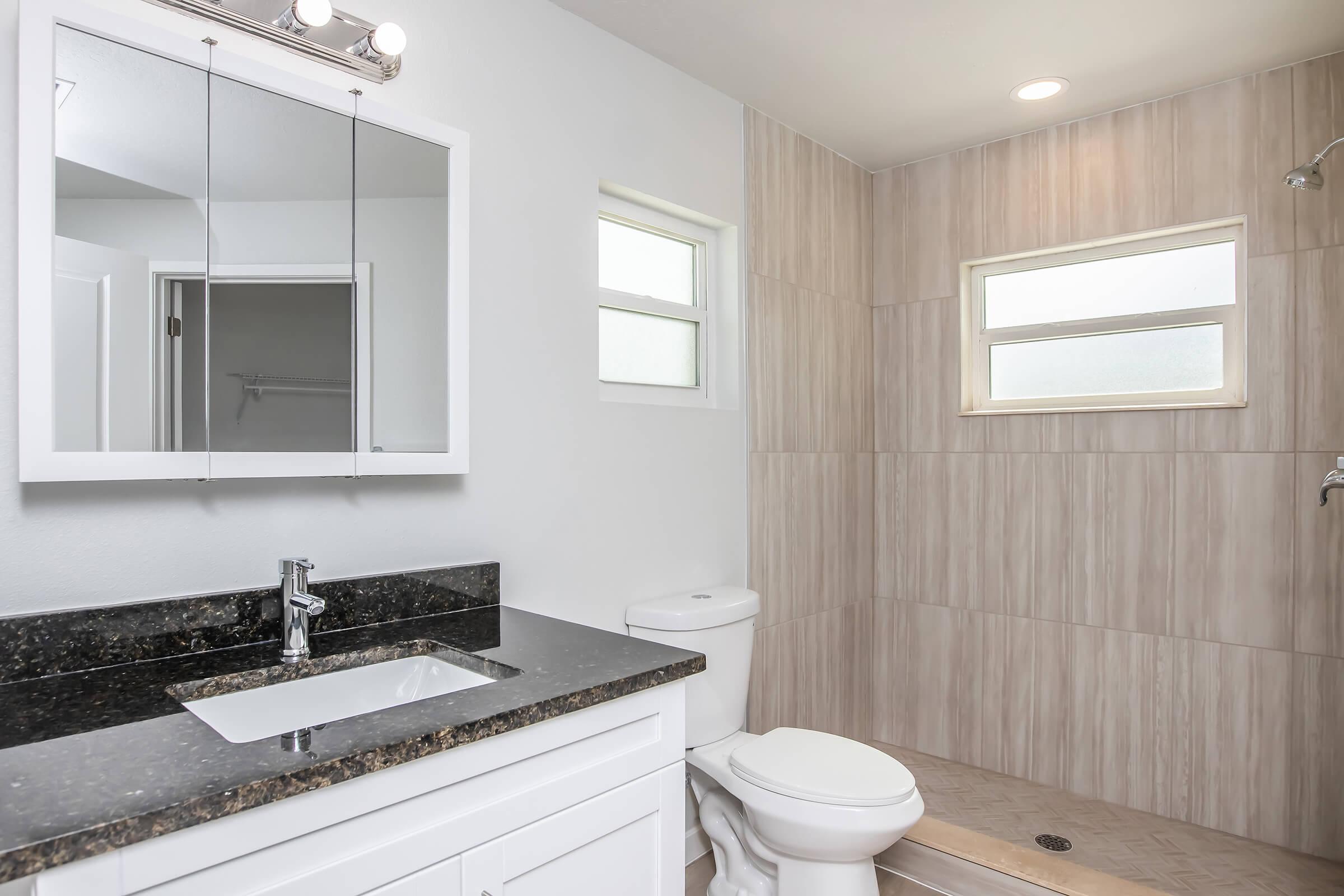 A modern bathroom featuring a white vanity with a dark granite countertop, a wall-mounted mirror, a white toilet, and a spacious shower with glass doors. Natural light enters through two small windows, and the walls are tiled in beige with a clean, contemporary design.