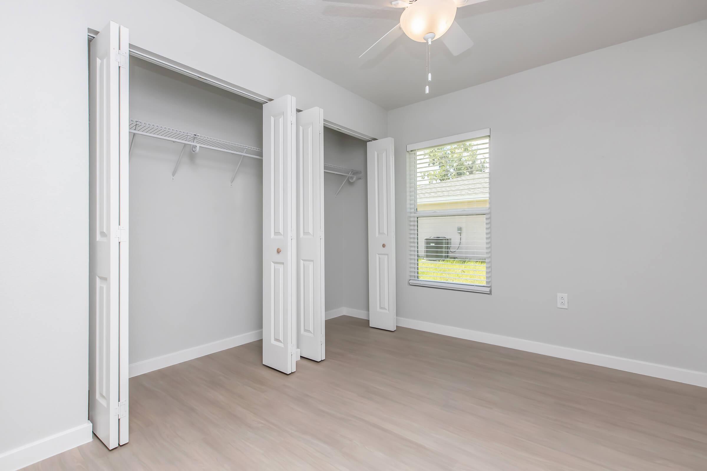 Empty room featuring light gray walls and light wood flooring. There are two white sliding closet doors open, revealing a simple wire closet system inside. A window with blinds lets in natural light, and a ceiling fan is visible. The overall ambiance is clean and minimalist.