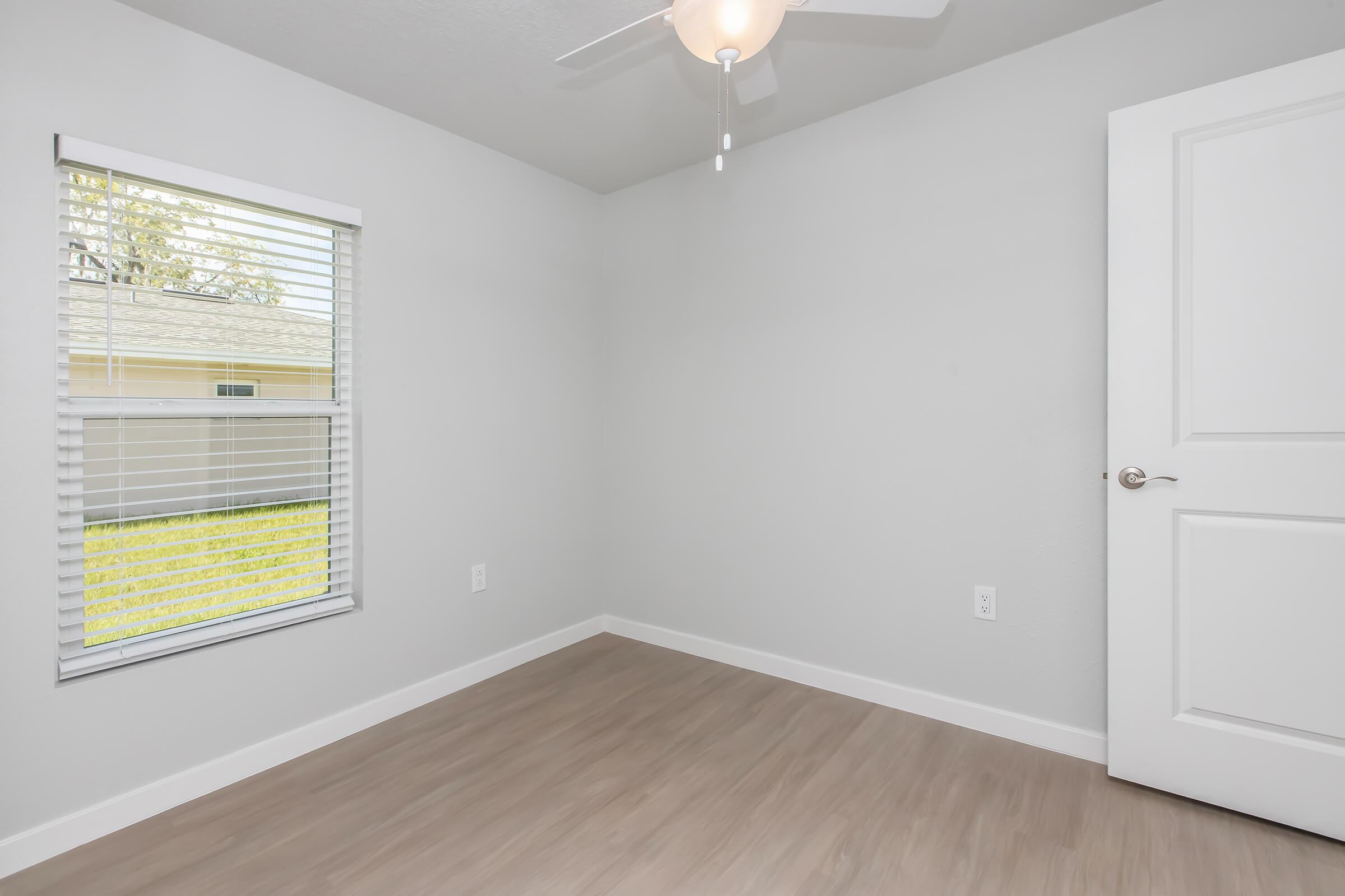 A clean, empty room with light gray walls and a light wood floor. There's a window with white blinds allowing natural light, and a ceiling fan is visible. A white door is positioned against one wall, adding to the simple and modern aesthetic of the space.