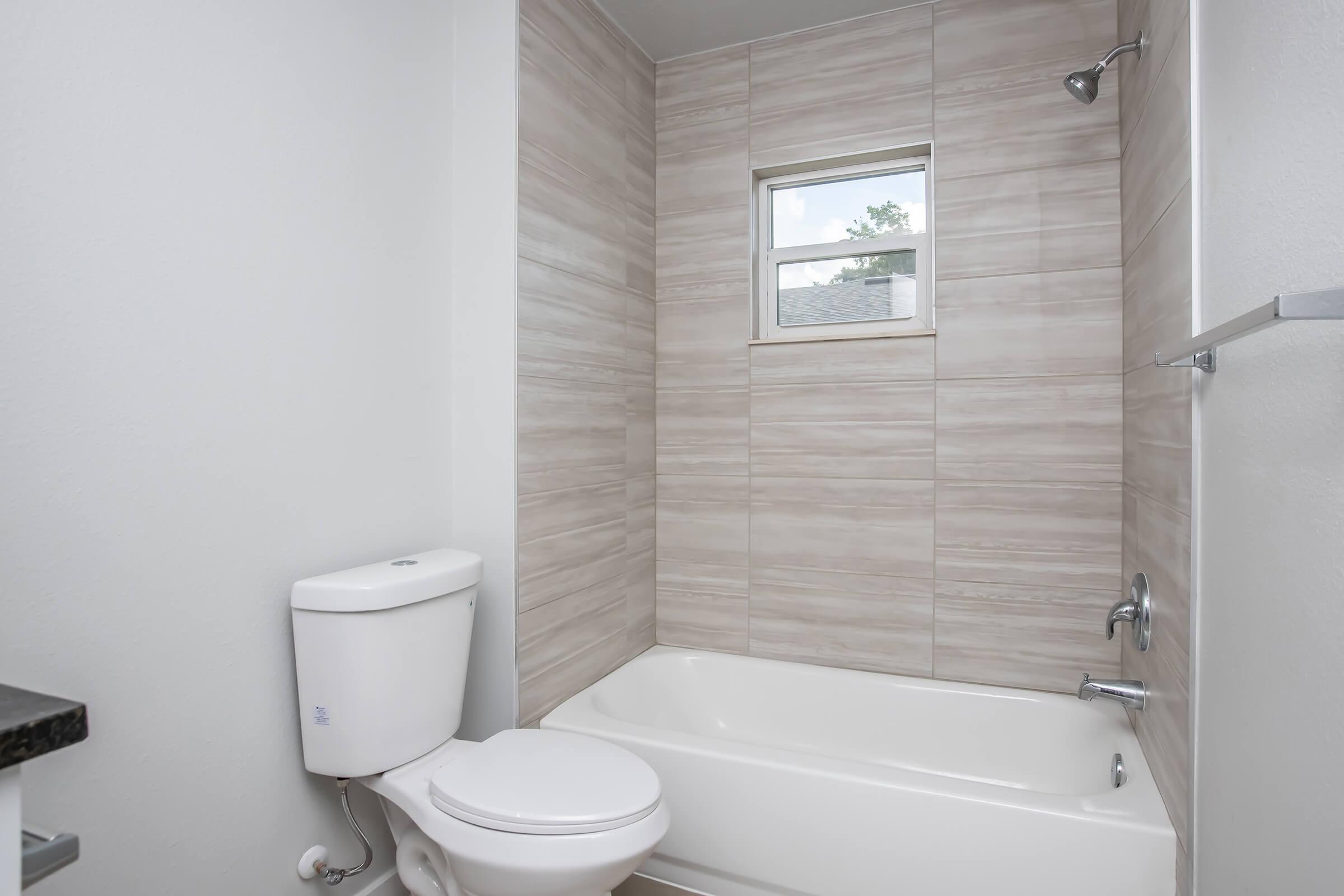A clean, modern bathroom featuring a bathtub with an adjacent shower, a white toilet, and light-colored tile walls. A small window allows natural light to enter the space, enhancing the airy feel. The overall design emphasizes a simple and functional aesthetic.