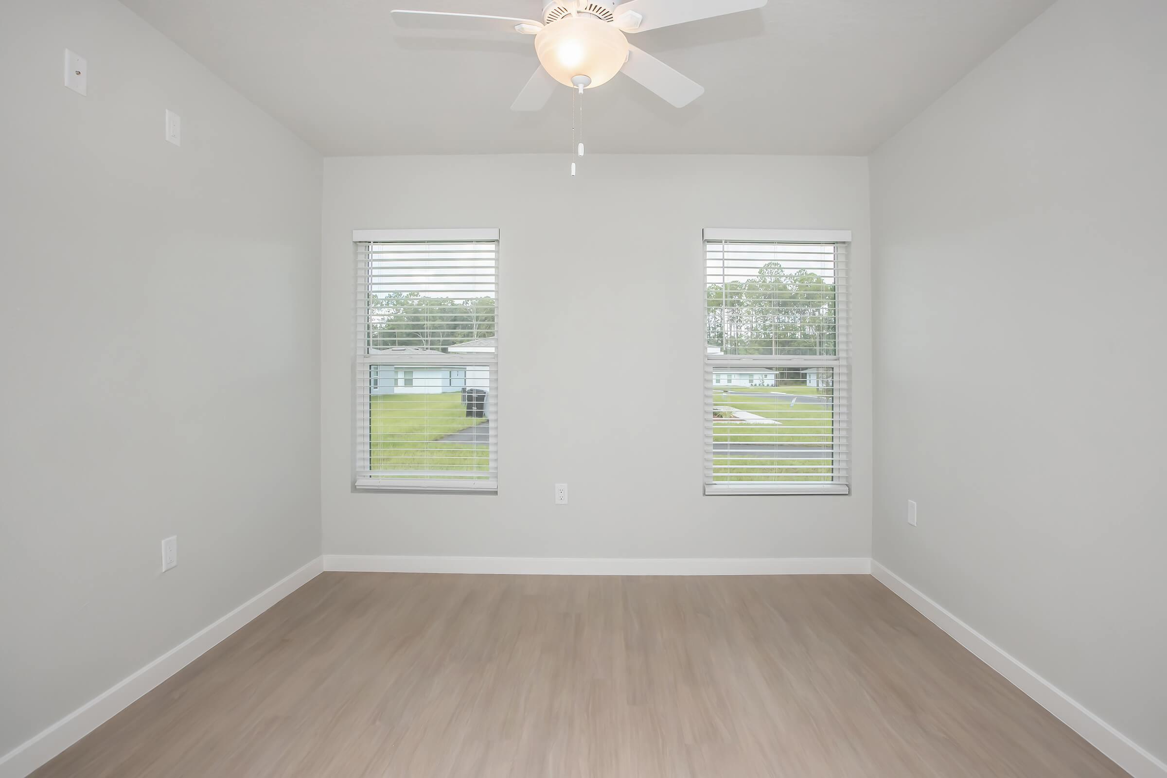 Bright, empty room with light gray walls and a ceiling fan. Two windows with blinds allow natural light in, and there is light wood flooring. The view outside shows a grassy area. The space is uncluttered and ready for furniture.