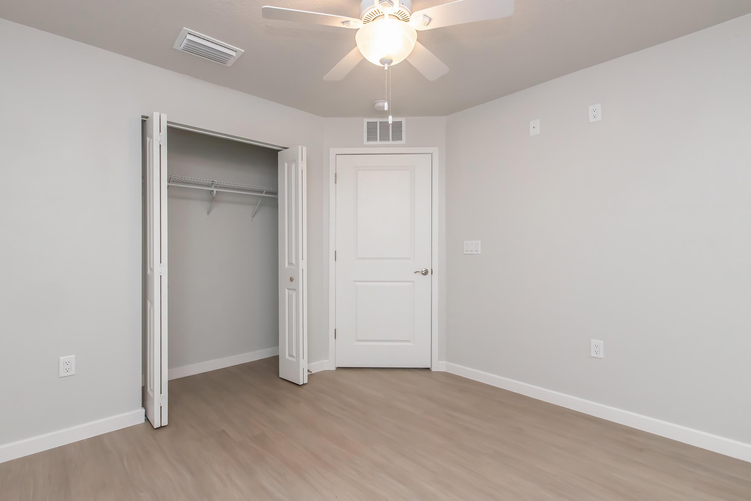 A well-lit, empty room featuring light gray walls and laminate flooring. There is a white ceiling fan, a closet with sliding doors on the left, and a doorway to another area. The room is spacious with minimal furnishings and natural light illuminating the space.