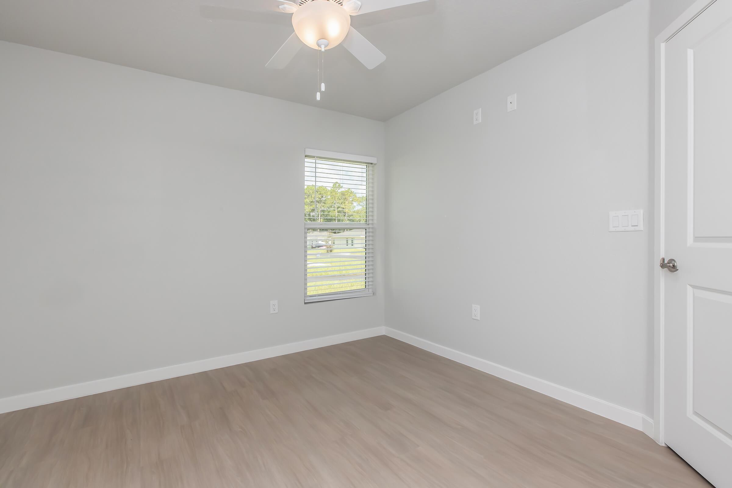 A clean, empty room with light gray walls and a ceiling fan. A window with blinds allows natural light to enter, illuminating the light wood flooring. The room has a door and two light switches on the adjacent wall. The overall atmosphere is bright and minimalistic.