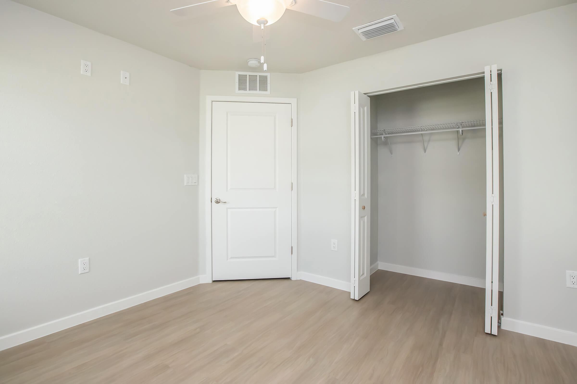 A clean, empty room featuring light-colored walls and flooring, with a ceiling fan. On one side, there is a closed white door leading to the hallway, and on the other, an open closet with a hanging rod and no shelves, highlighting spaciousness and simplicity in design.
