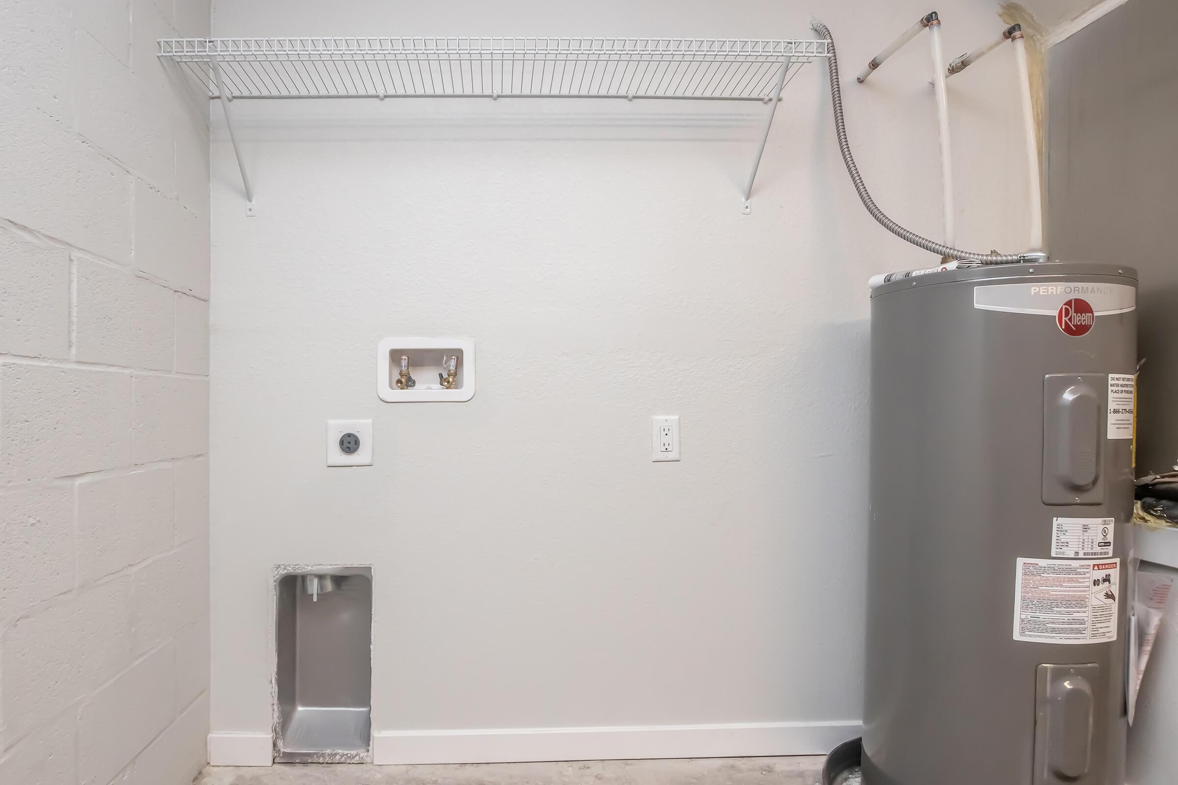 A laundry room with a gray wall, a wire shelf mounted above, and a gray water heater on the right. There are plumbing and electrical connections visible on the wall, with an empty space on the floor designed for a washer or dryer installation. The area is well-lit and uncluttered.