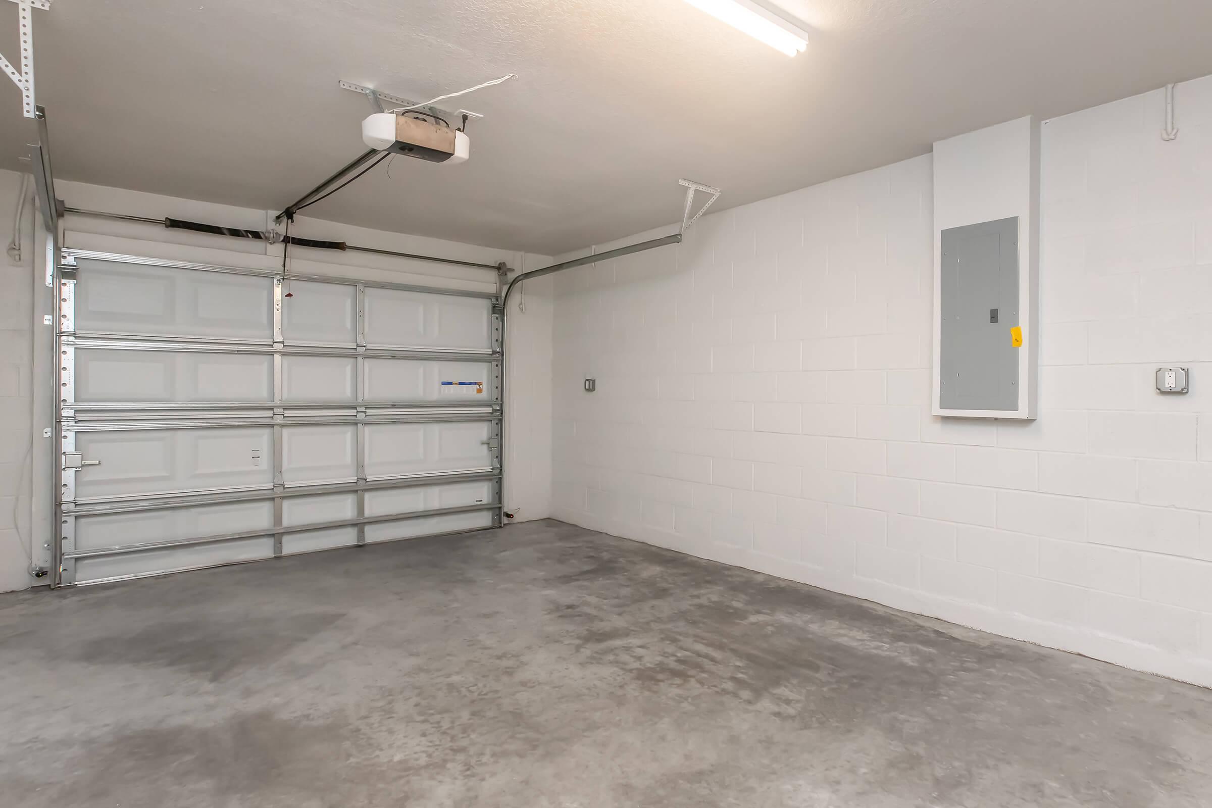 Interior view of a clean and empty garage featuring a white wall, a concrete floor, a closed roll-up garage door, and an electrical panel on the wall. Bright overhead lighting illuminates the space.