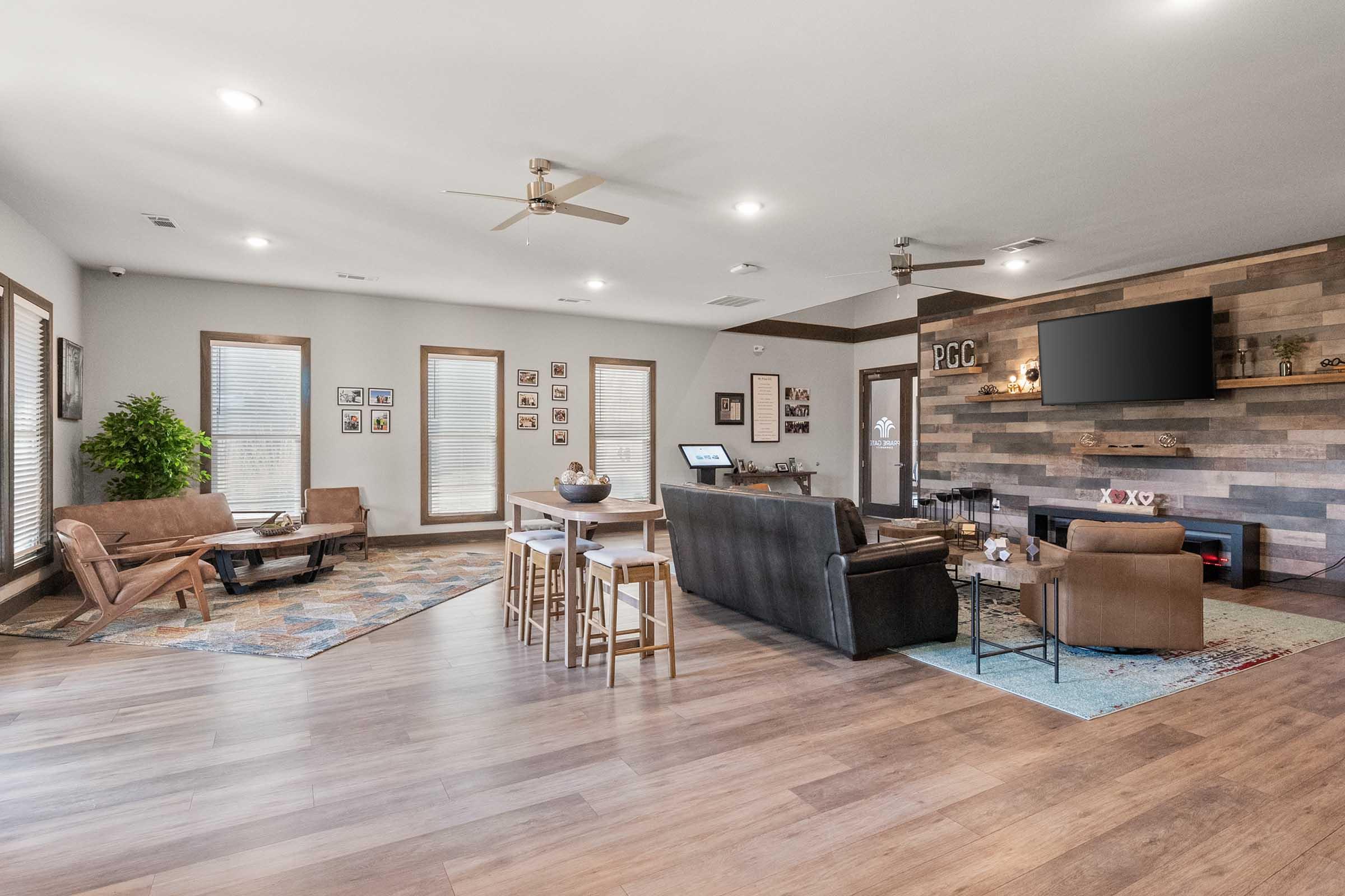 A spacious living room featuring wooden floors, a comfortable seating area with brown leather chairs and a sofa, a coffee table, and a dining table. The walls are adorned with framed pictures, and a modern fireplace is visible with a large TV above it. Large windows allow natural light to fill the space, and a potted plant adds a touch of greenery.