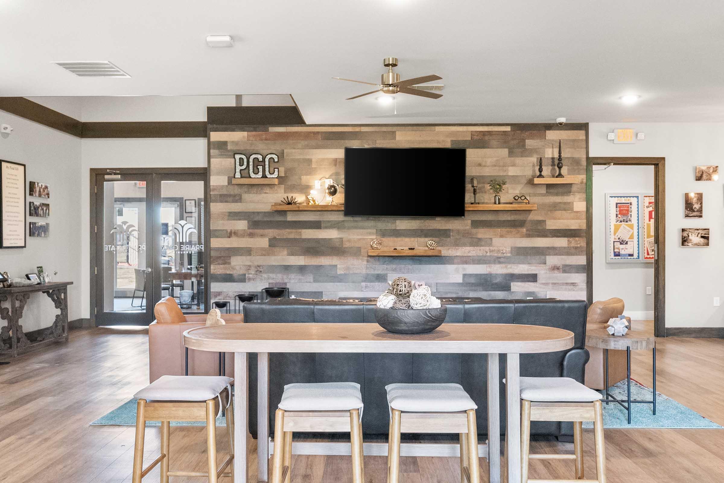 A modern lounge area featuring a wooden accent wall with a TV mounted on it. There is a table with seating in front, and a decorative bowl on the table. The space is well-lit with a ceiling fan, and there are blue and neutral color accents throughout.