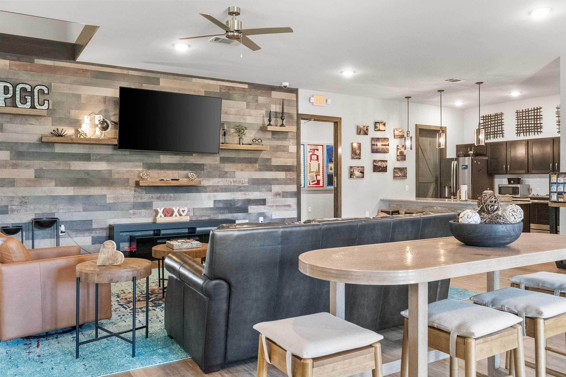 A modern living area with a mix of seating options, including a black leather couch and an orange armchair. There's a wooden dining table with simple chairs, and a large flat-screen TV mounted on a wood-paneled wall. The space features a neutral color palette with decorative elements, including wall art and a rug, creating a cozy atmosphere. The kitchen area is visible in the background with dark cabinetry and modern appliances.