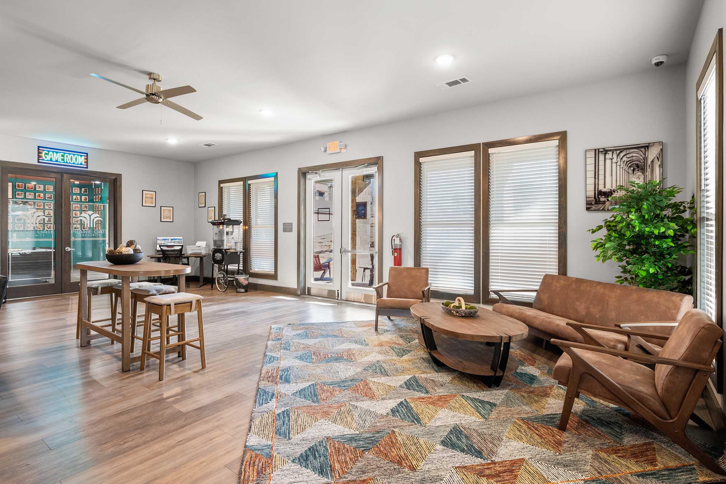 A modern, welcoming common area featuring a brown leather sofa, two armchairs, a round coffee table, and a wooden high-top table with stools. The floor is wood, and a colorful geometric area rug adds warmth. Large windows allow natural light, and there are plants and decorative elements around the room. A game room is visible through the glass door in the background.