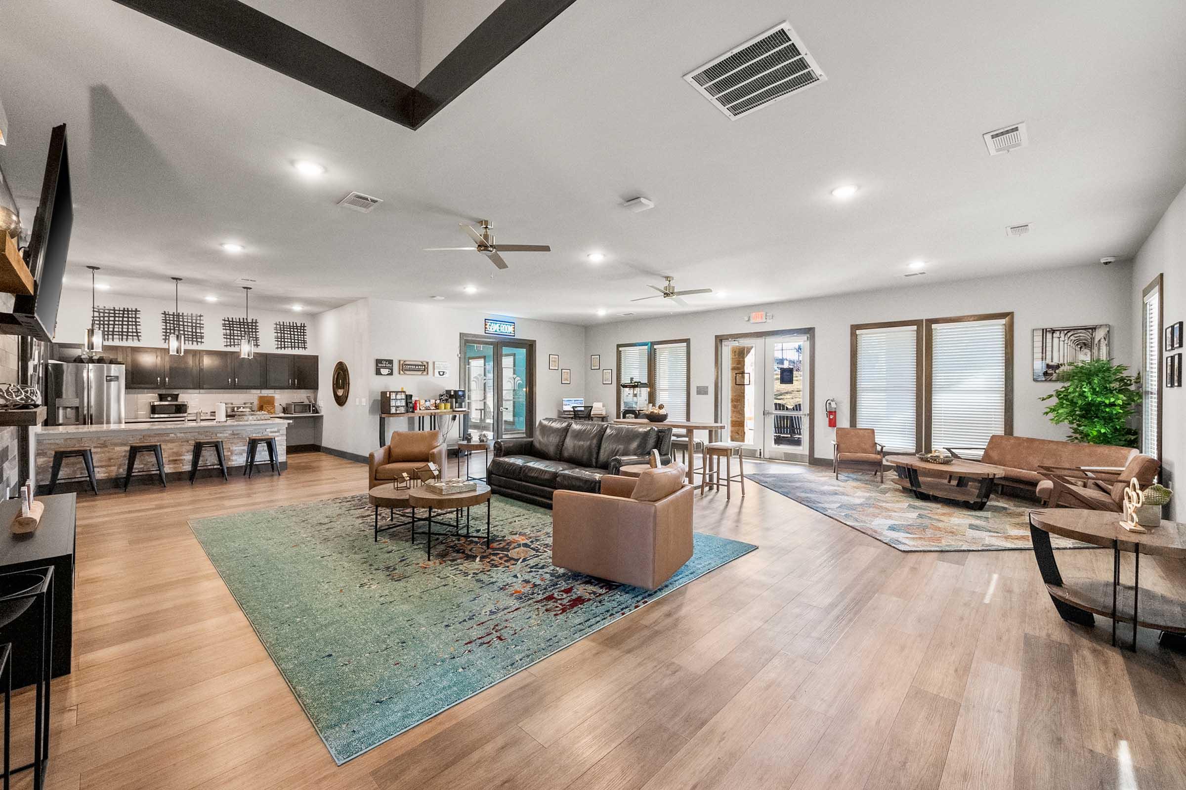 A spacious, modern living room area featuring a dark leather sofa, two tan armchairs, a coffee table, and a large area rug. The room has wooden flooring, large windows, and is furnished with artwork and plants. A kitchen area with bar seating is visible in the background.