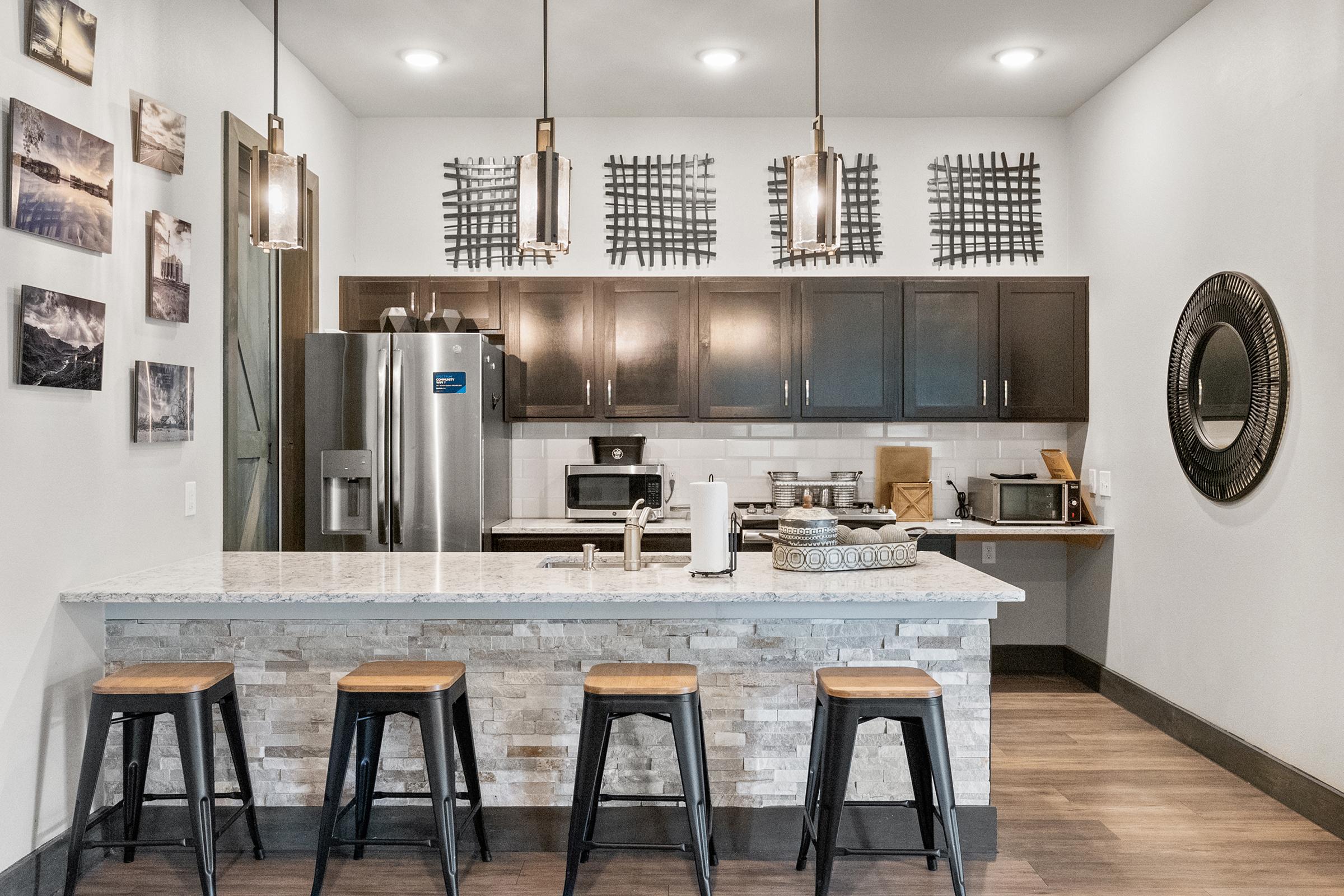 Modern kitchen interior featuring dark cabinetry, a white countertop with stone accents, three wooden bar stools, stainless steel appliances, decorative wall art, and stylish pendant lighting.