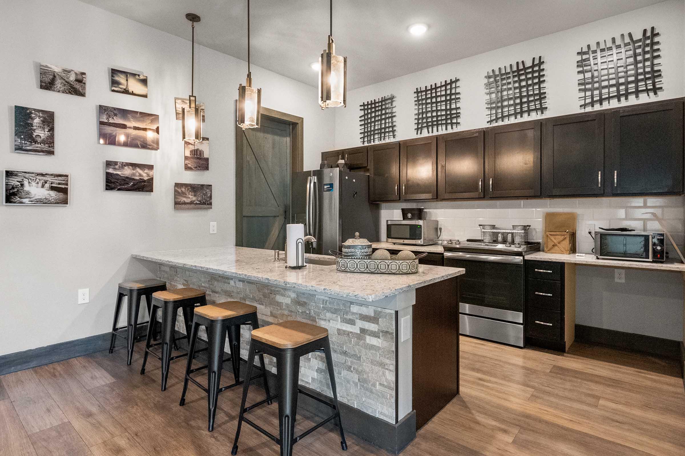 A modern kitchen featuring a large island with bar stools, dark cabinets, stainless steel appliances, and decorative wall art. The space has warm wooden flooring and industrial-style pendant lights hanging above the island.