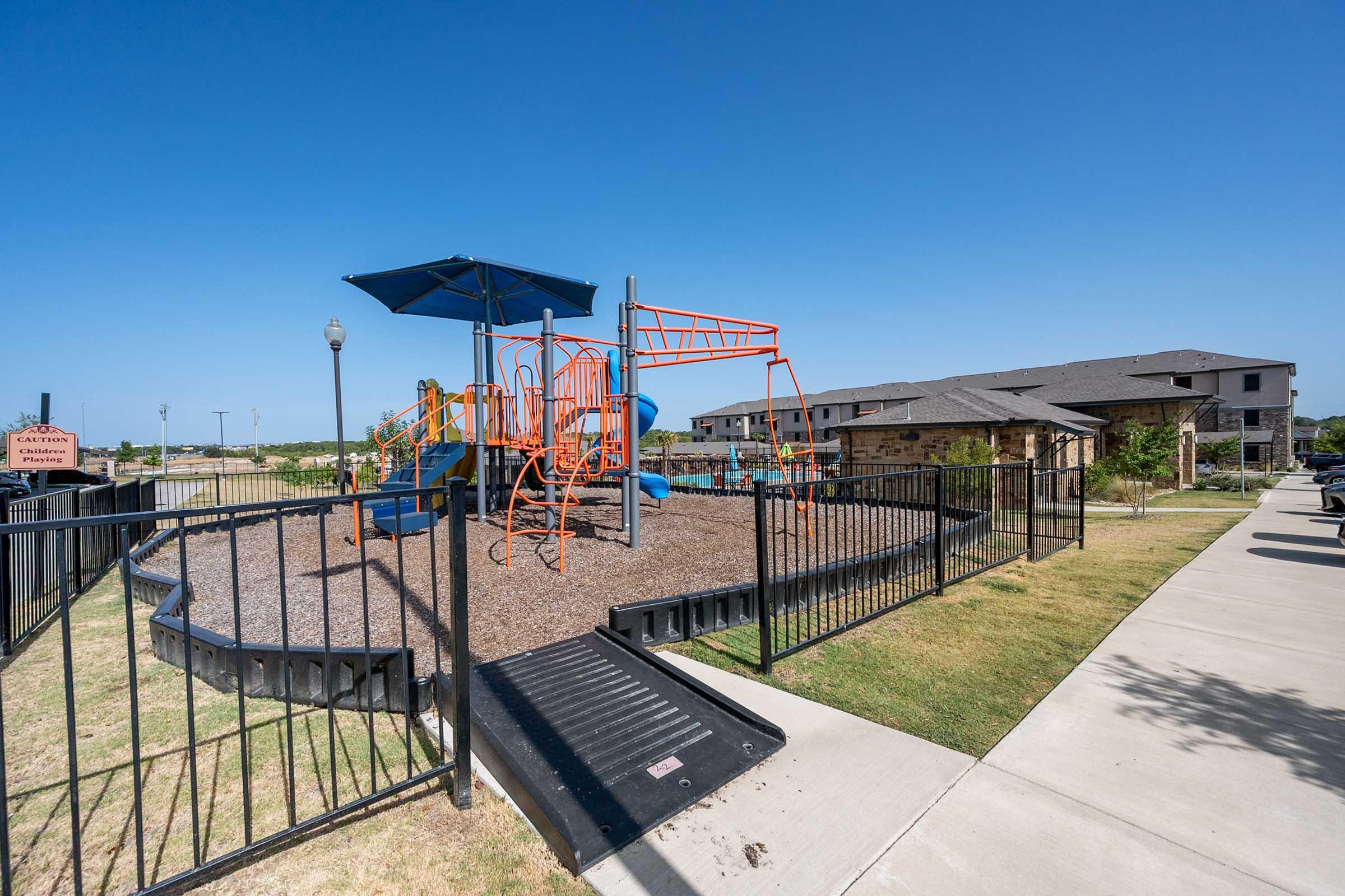 A playground structure with orange equipment, including swings and climbing features, is surrounded by a black fence. A blue shade canopy is positioned above the playground. In the background, there are residential buildings and a clear blue sky. A concrete pathway runs alongside the playground.