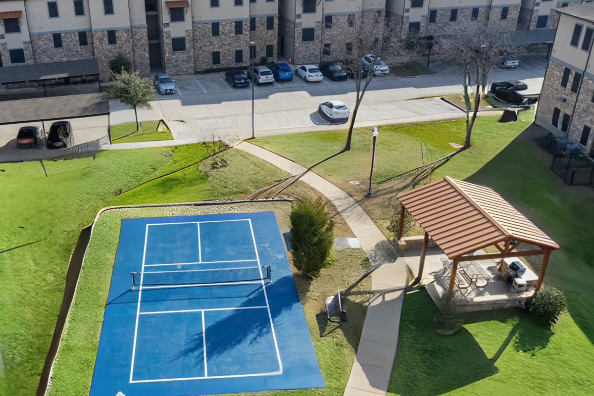 Aerial view of a tennis court with a blue surface, surrounded by green grass and a walking path. A gazebo with seating is visible nearby, along with parked cars in the background and a building in the distance.