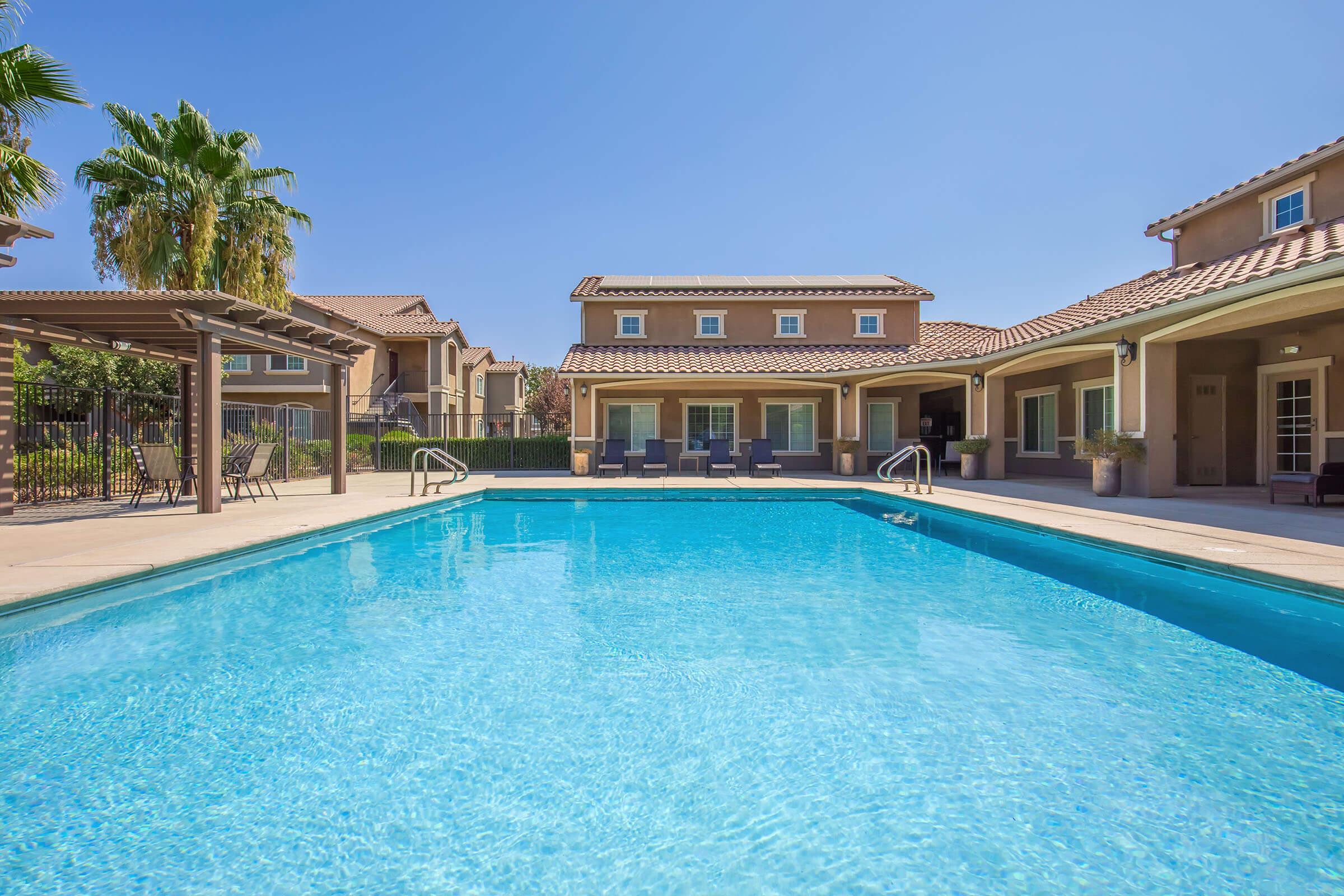 A clear, blue swimming pool surrounded by a patio area. In the background, there are tan-colored residential buildings with tiled roofs, palm trees, and green landscaping. The scene is bright and sunny, creating a relaxing outdoor atmosphere.