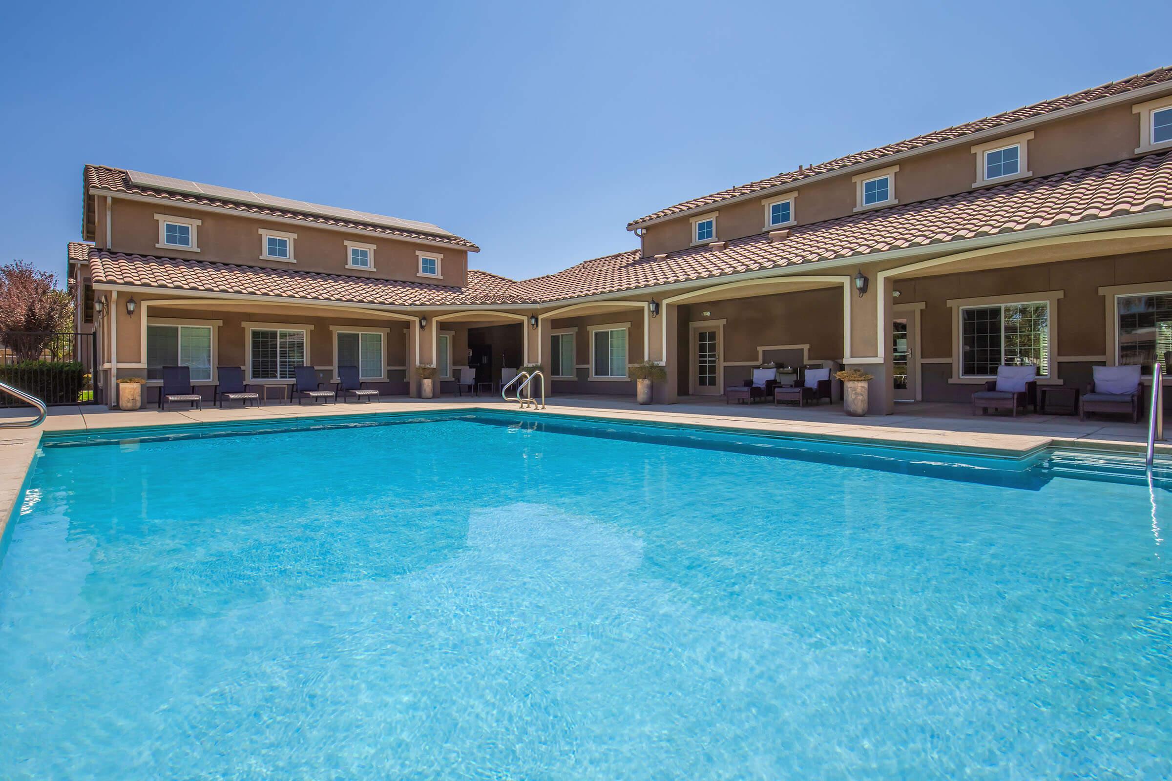 A clear blue swimming pool in a well-maintained backyard, surrounded by a spacious patio with seating areas. In the background, a two-story house featuring large windows and a tiled roof is visible. The scene is set under a bright blue sky, suggesting a sunny day.