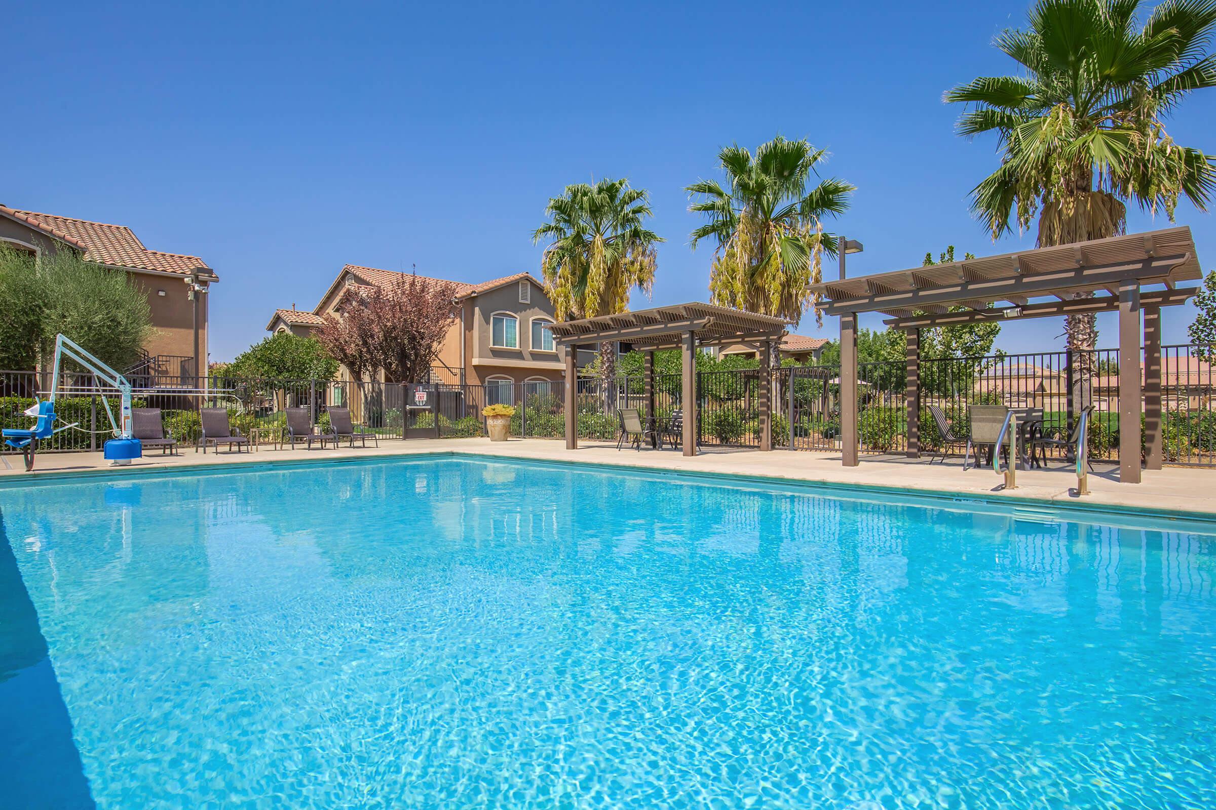 A clear blue swimming pool surrounded by palm trees and lounge chairs. There are pergolas and landscaped areas in the background, with residential buildings visible. The sky is sunny and bright, creating a warm, inviting atmosphere.