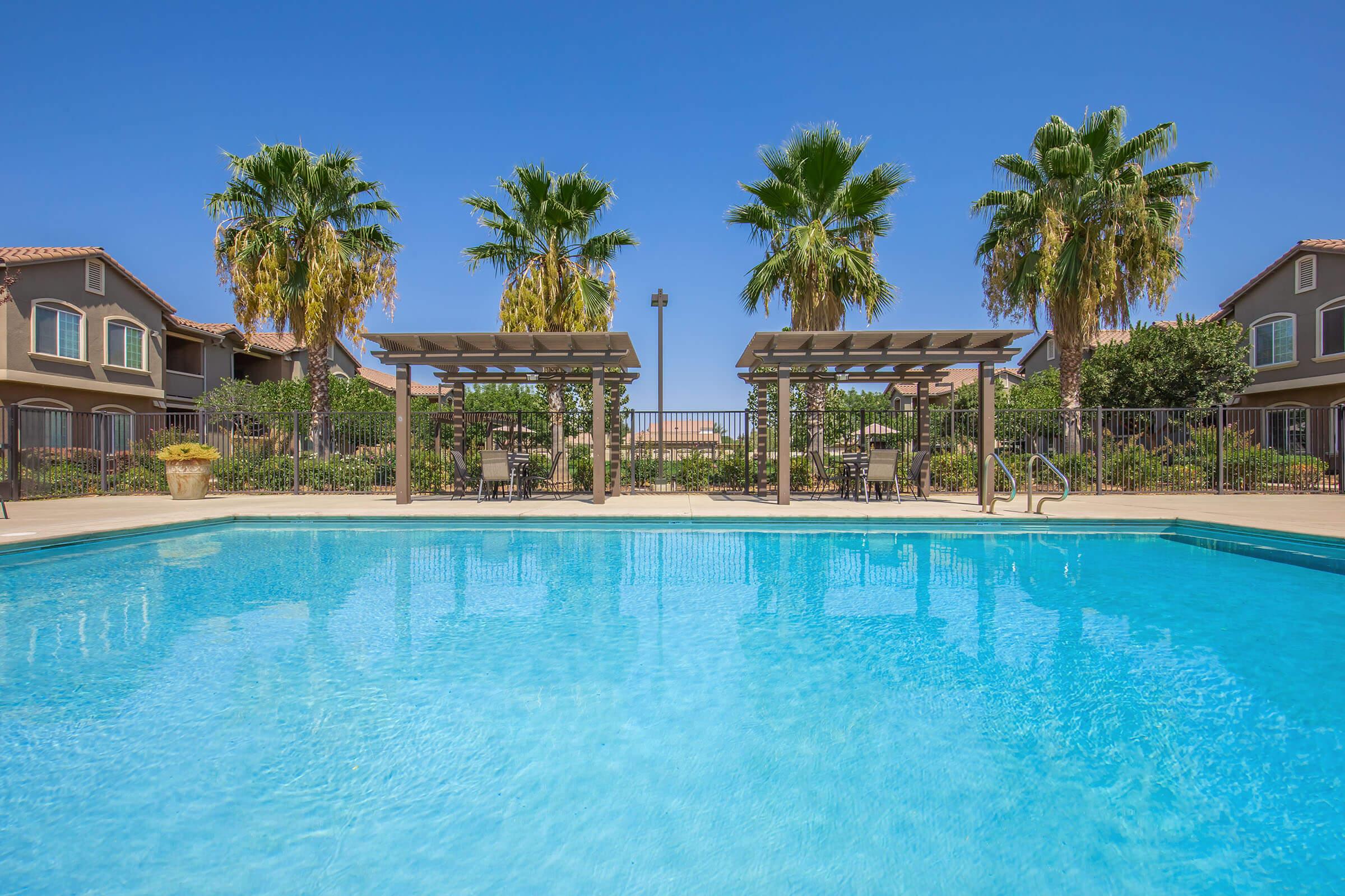 A clear blue swimming pool surrounded by lush greenery and palm trees, with shaded seating areas under wooden pergolas. In the background, residential buildings are visible against a bright blue sky. The scene conveys a relaxing outdoor space ideal for leisure and recreation.
