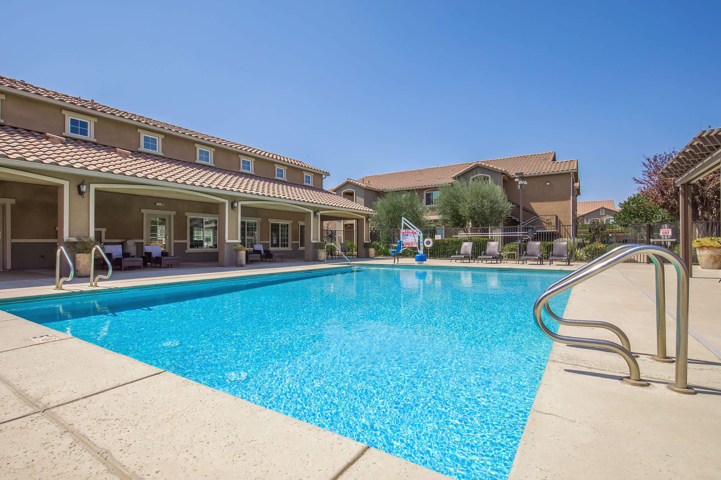 A sunny outdoor pool area featuring a clear blue pool with a set of stairs for access. Surrounding the pool are lounge chairs and shaded seating areas. In the background, there are residential buildings with balconies and landscaped greenery. The sky is clear and blue, creating a relaxing atmosphere.