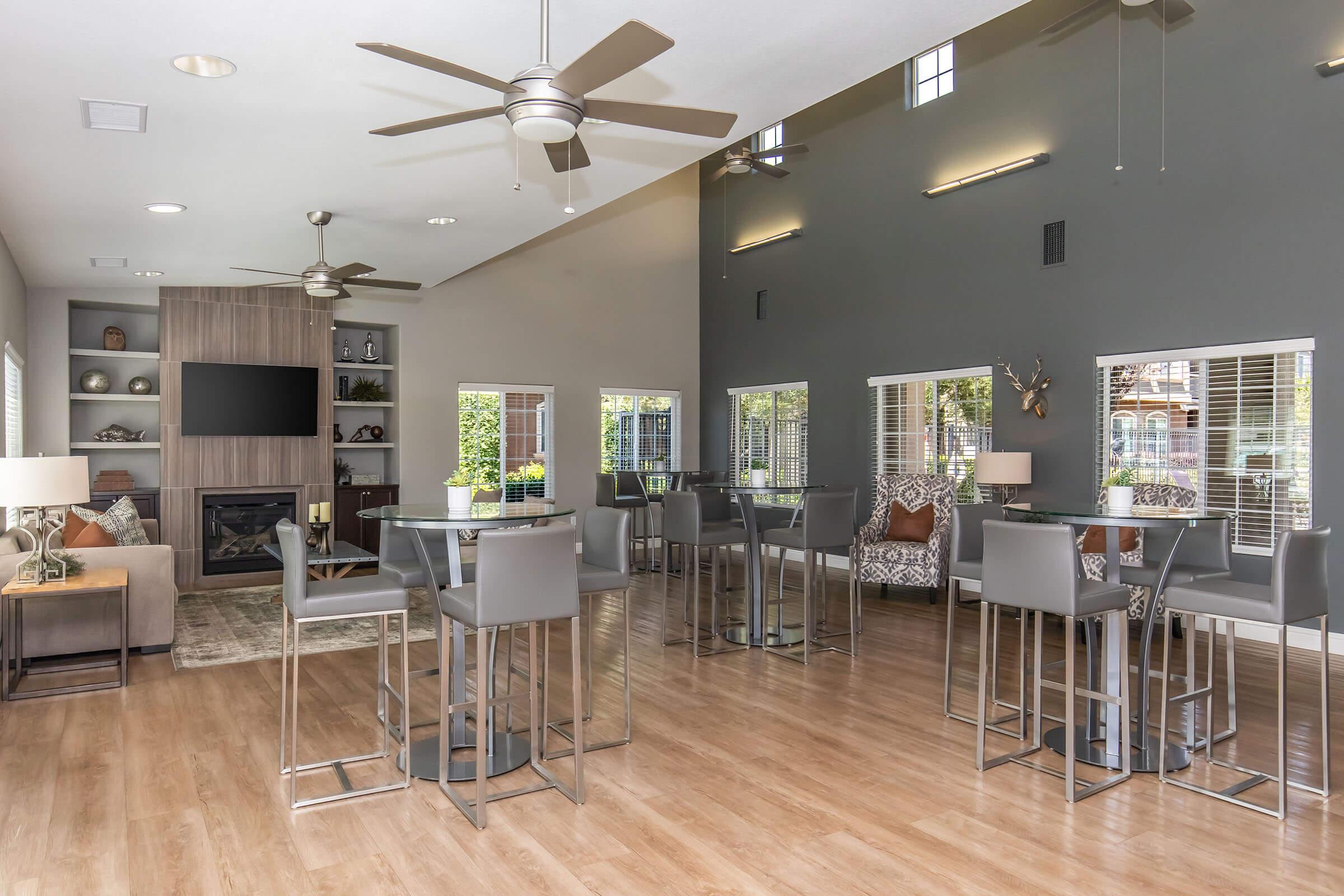 A modern lounge area featuring stylish furniture, including high-top tables and chairs. The space is well-lit with large windows, a flat-screen TV mounted on the wall, and a cozy fireplace. The decor includes neutral tones and decorative elements, creating a welcoming atmosphere.