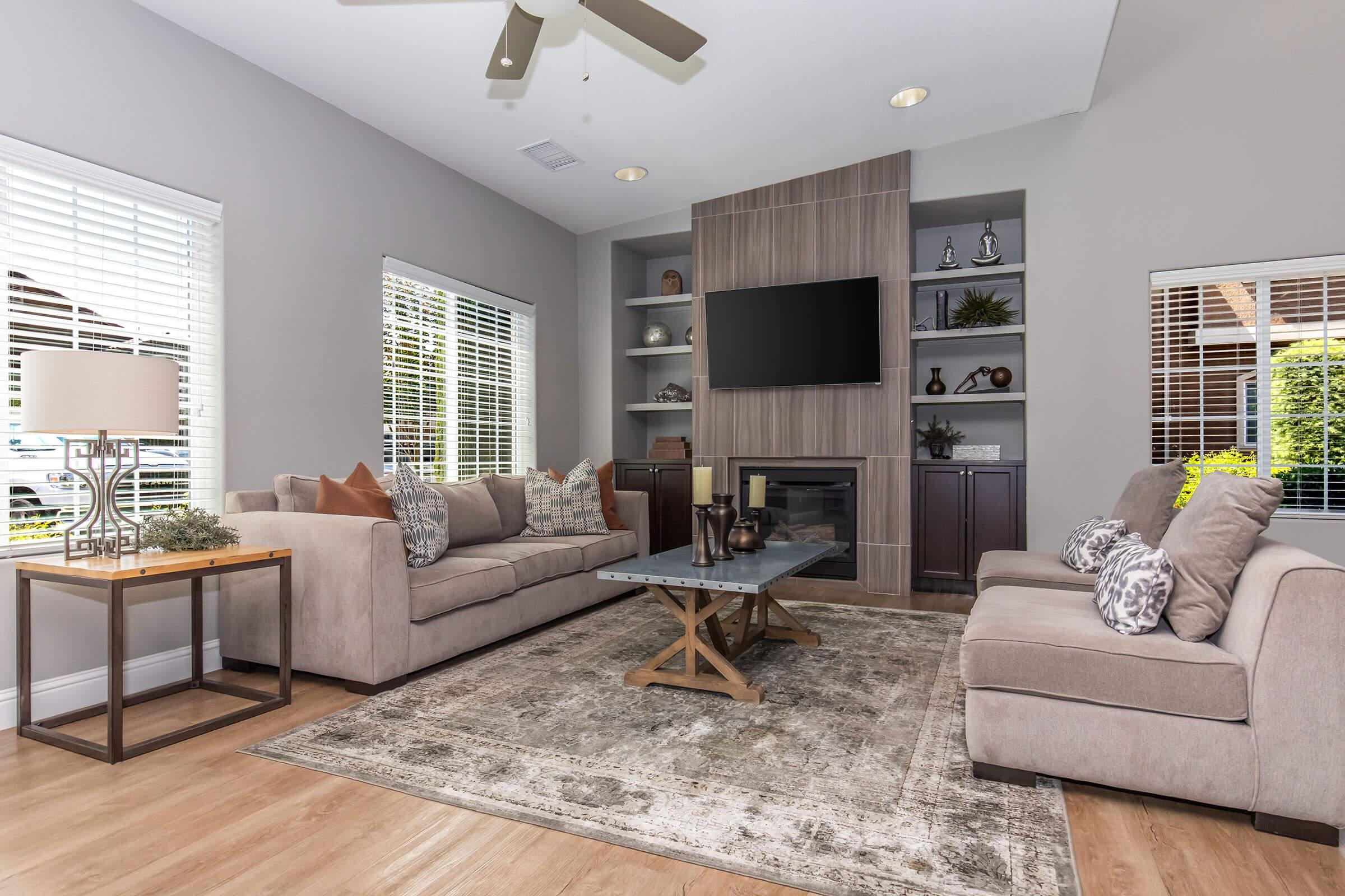A modern living room featuring two beige sofas, a wooden coffee table with a geometric design, and a decorative area rug. A wall-mounted television is above a contemporary fireplace, and there are large windows with blinds allowing natural light. A lamp and decorative items are on the side table.
