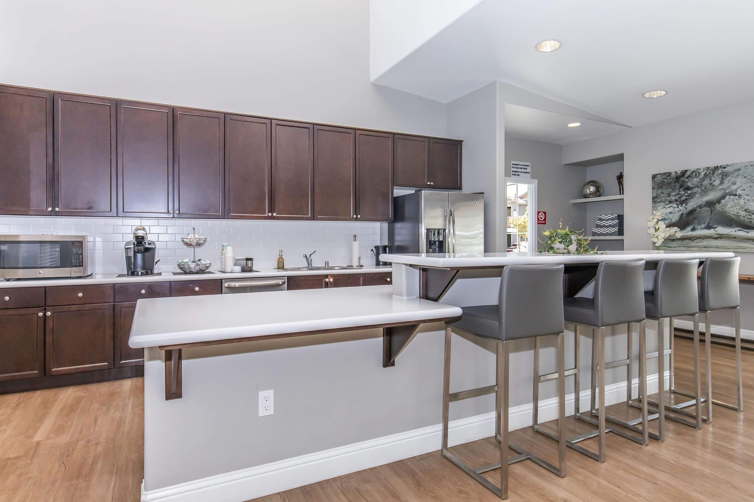 Modern kitchen featuring dark wood cabinets, stainless steel appliances, and a sleek countertop. A dining area with high-backed stools sits in front of the counter, and light-colored walls provide a bright ambiance. The space is clean and contemporary, with decorative elements adding a touch of elegance.