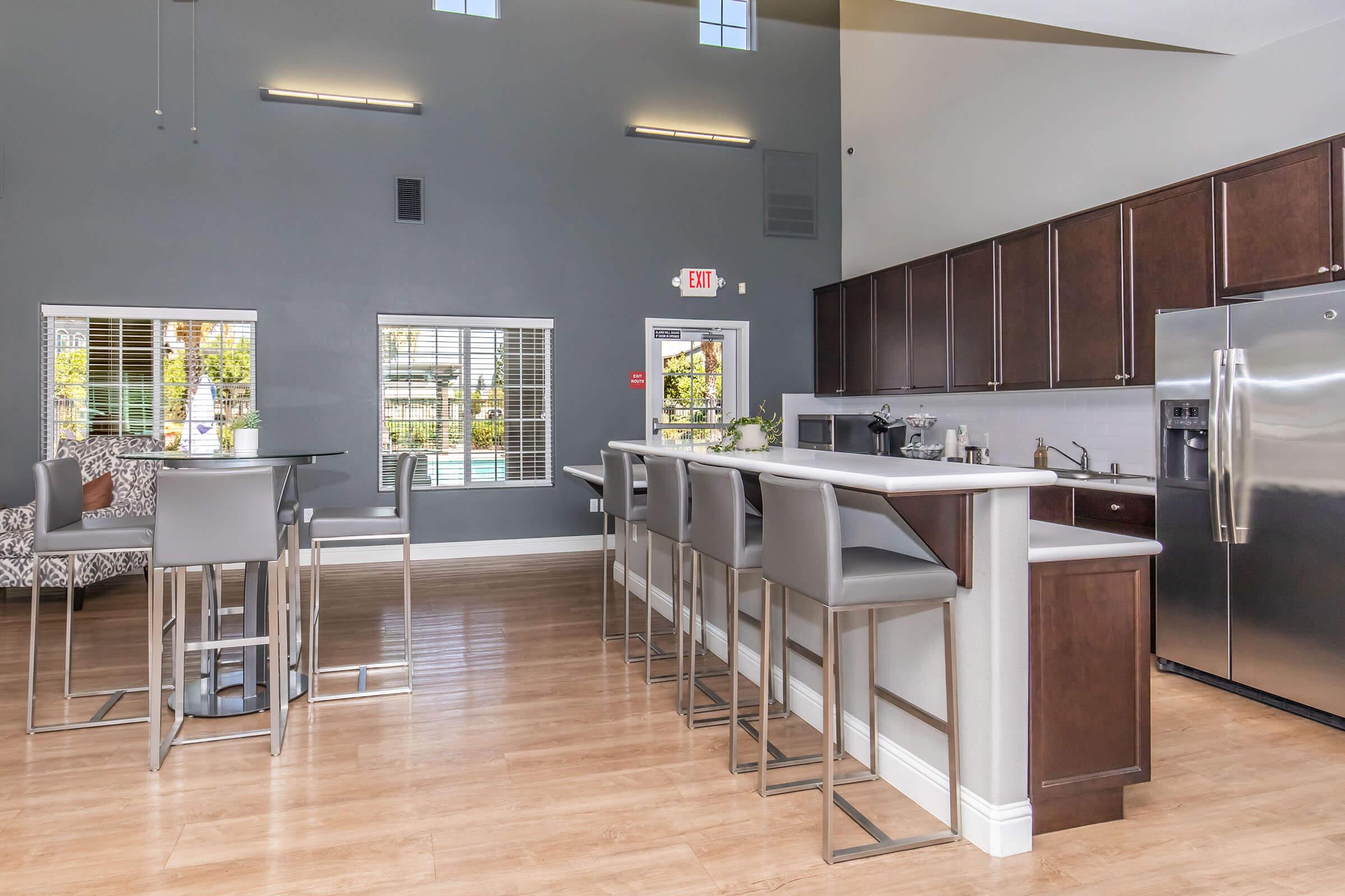 A modern kitchen and dining area with a large island and bar seating. The kitchen features sleek cabinetry and stainless steel appliances. Large windows provide natural light, and there is a view of a patio outside. The flooring is light wood, creating a welcoming atmosphere.