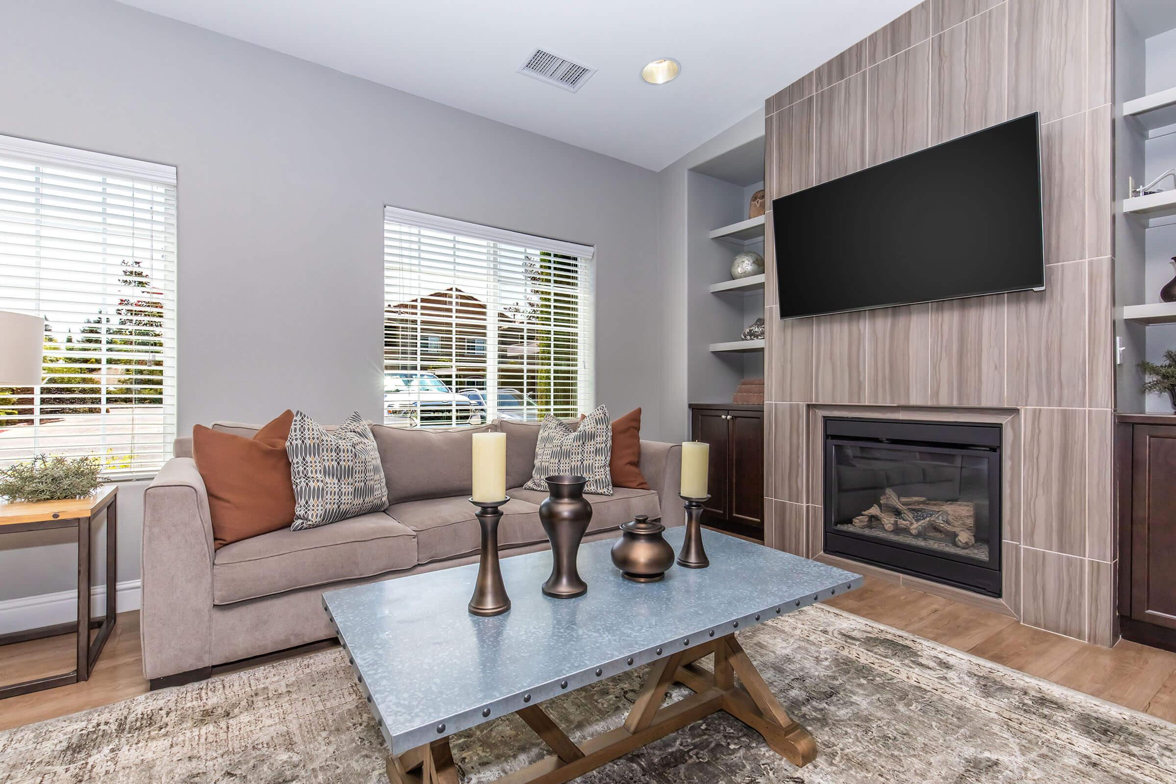 A modern living room featuring a light gray sofa with orange and patterned pillows. A coffee table made of metal and wood sits in front, adorned with decorative candles and vases. There are two large windows with white blinds, and a built-in fireplace with a TV mounted above. The walls are light gray with wooden accents.