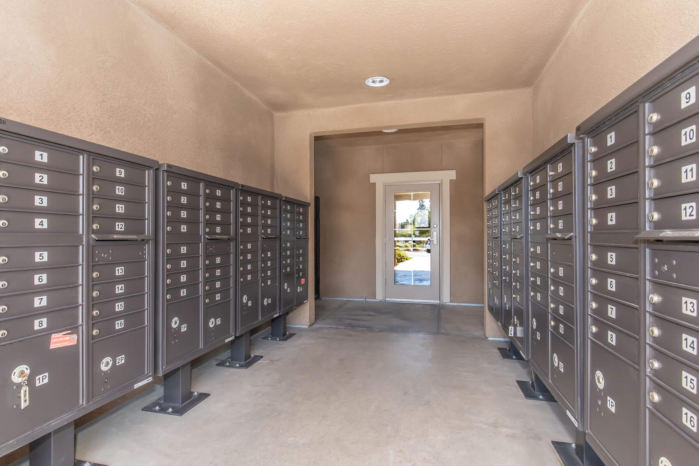 A corridor lined with dark gray mailboxes, featuring multiple compartments for mail storage on either side. At the end of the corridor, there is a door leading to the outside, framed by light-colored walls and a ceiling. The space is well-lit and tidy, with a concrete floor.