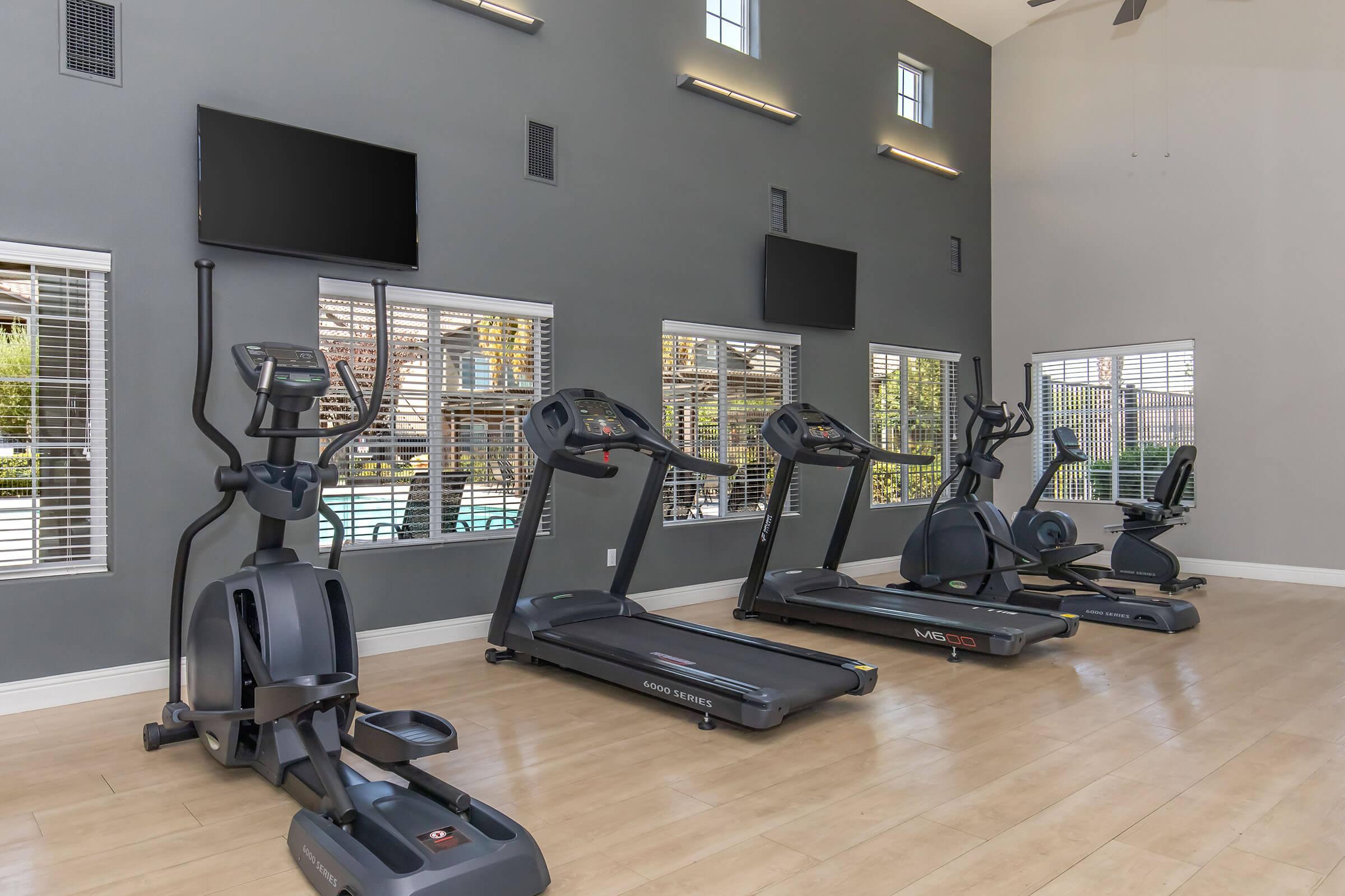 A modern gym interior featuring several exercise machines, including treadmills and an elliptical, positioned against a gray wall with large windows letting in natural light. Television screens are mounted above the machines, and a pool area is visible outside through the windows.