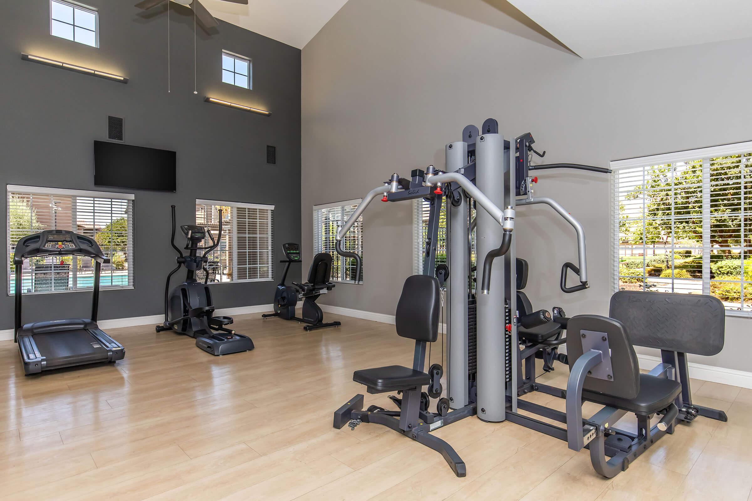 A modern gym interior featuring various exercise equipment including a multi-station weight machine, treadmill, and elliptical. The space has large windows allowing natural light, with a neutral color scheme and wooden flooring, creating a bright and inviting atmosphere for workouts.