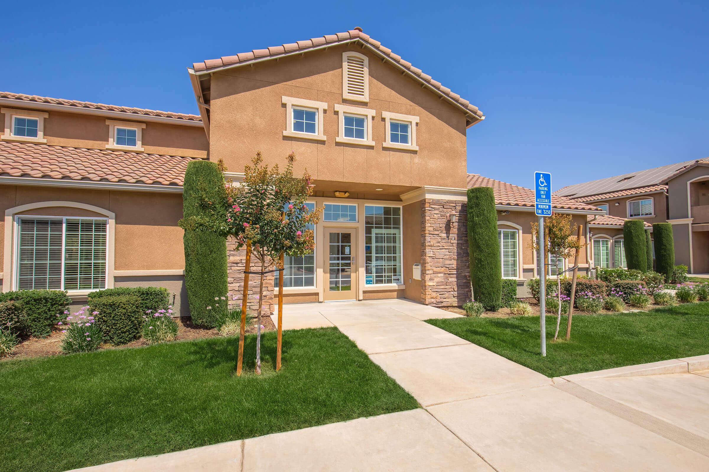 A modern residential building with a tan exterior and stone accents, featuring a welcoming entrance with large windows. The surrounding landscape includes neatly trimmed grass, small trees, and flowering shrubs, with a clear blue sky above and a designated parking space for individuals with disabilities nearby.