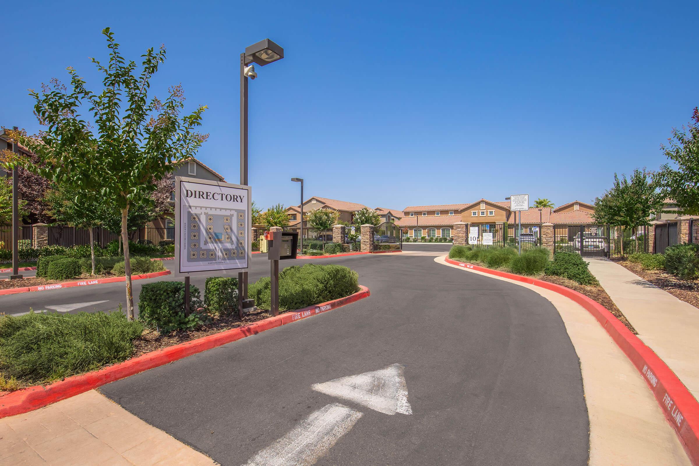 A view of a landscaped entrance to a community or complex, featuring a directory sign on the left, a winding road with a white arrow directing traffic, and buildings in the background under a clear blue sky. Green shrubs line the edges of the road.