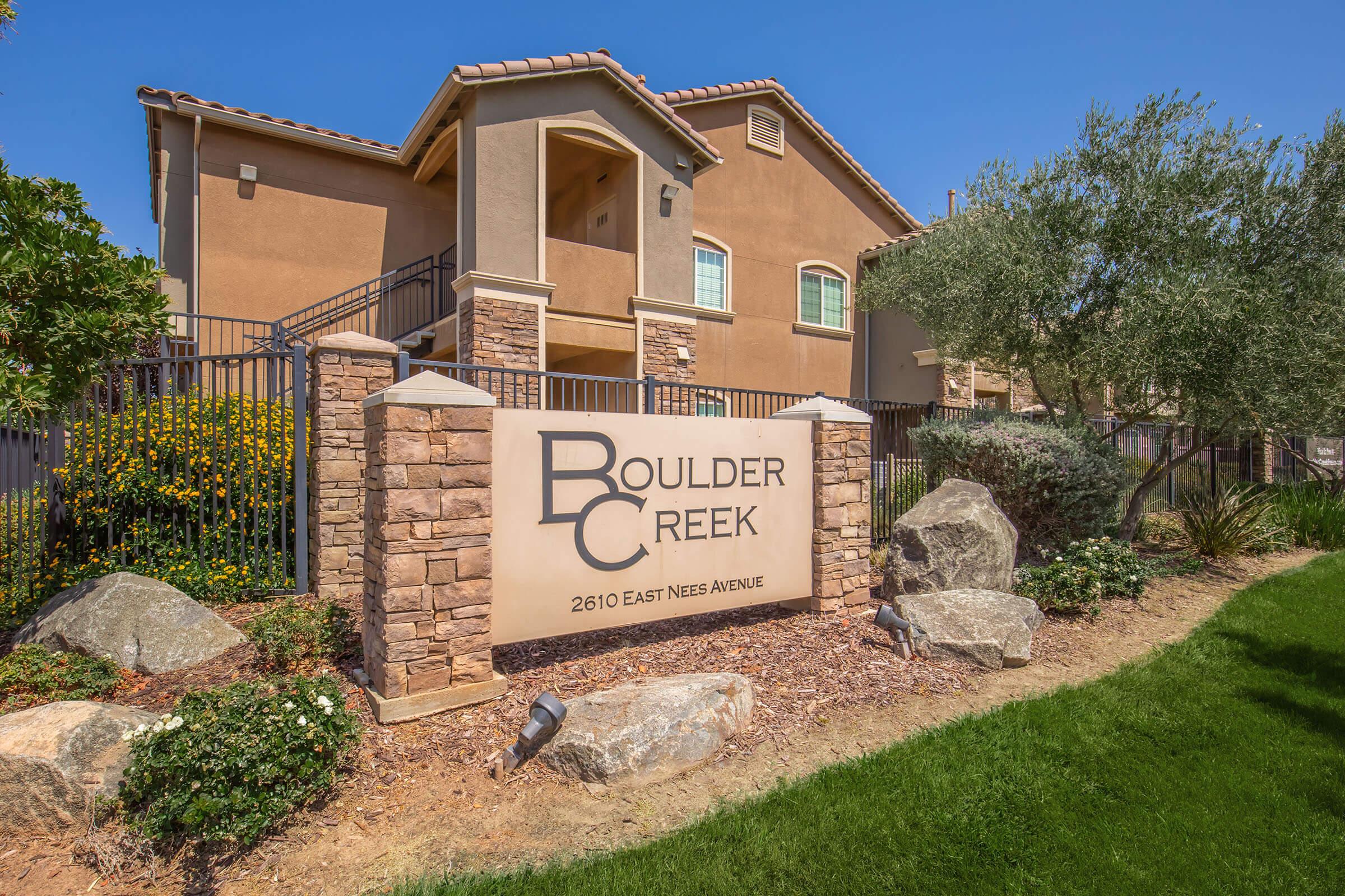 Sign for Boulder Creek with the address 2610 East Nees Avenue, surrounded by landscaped greenery and rocks, featuring a residential building in the background under a clear blue sky.