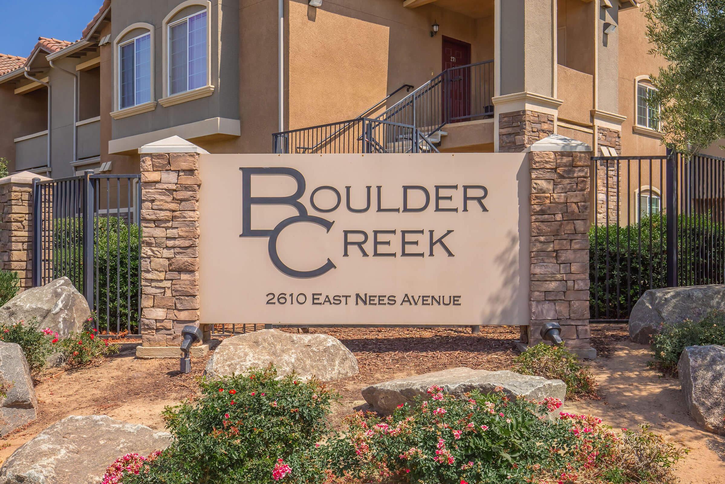 Sign for Boulder Creek located at 2610 East Nees Avenue, featuring stylized text with "BC" prominently displayed. Surrounding the sign are landscaped rocks and flowers, with a multi-story building visible in the background.