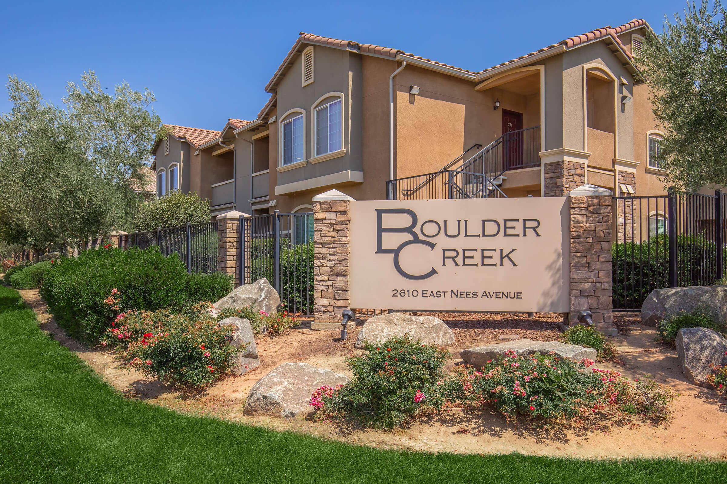 A stone sign for Boulder Creek Apartments featuring the name prominently displayed, surrounded by landscaped greenery, flowers, and rocks. The building is a tan color with architectural details, set against a clear blue sky.