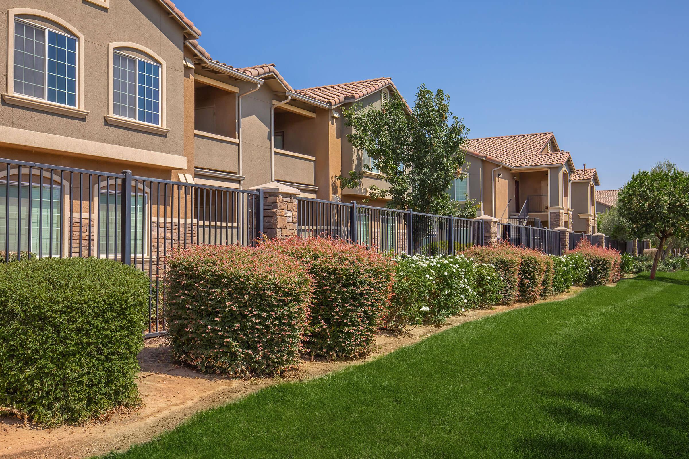 A landscaped area featuring well-maintained green grass, colorful bushes, and a fence. In the background, there are two-story residential buildings with balconies and large windows, under a clear blue sky. The scene conveys a calm, suburban environment.