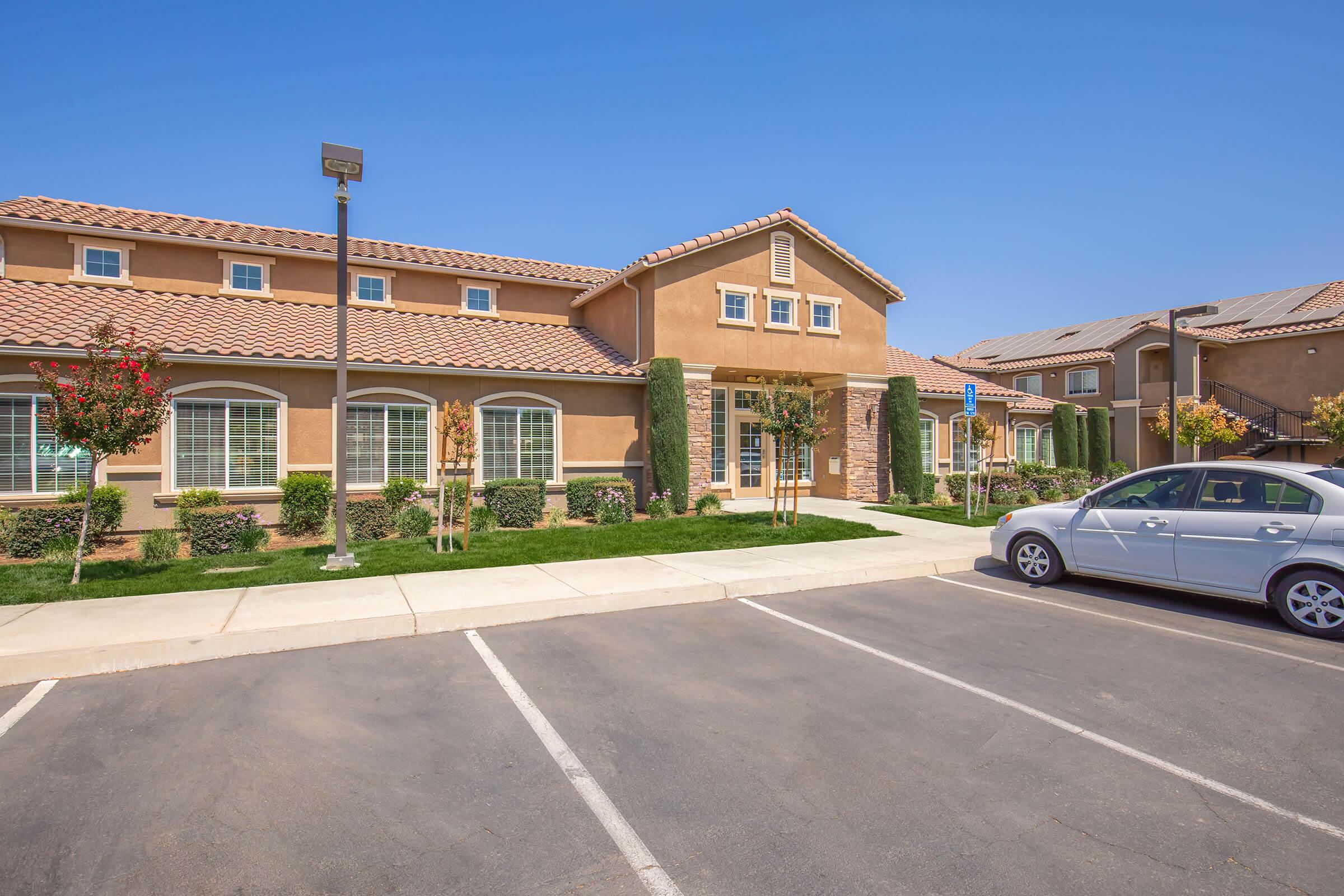 A well-maintained residential building with a beige exterior and red tile roof. The front features landscaped greenery and parking spaces. A silver car is parked in the lot, and the sky is clear and blue. Trees and shrubs enhance the property’s appeal.