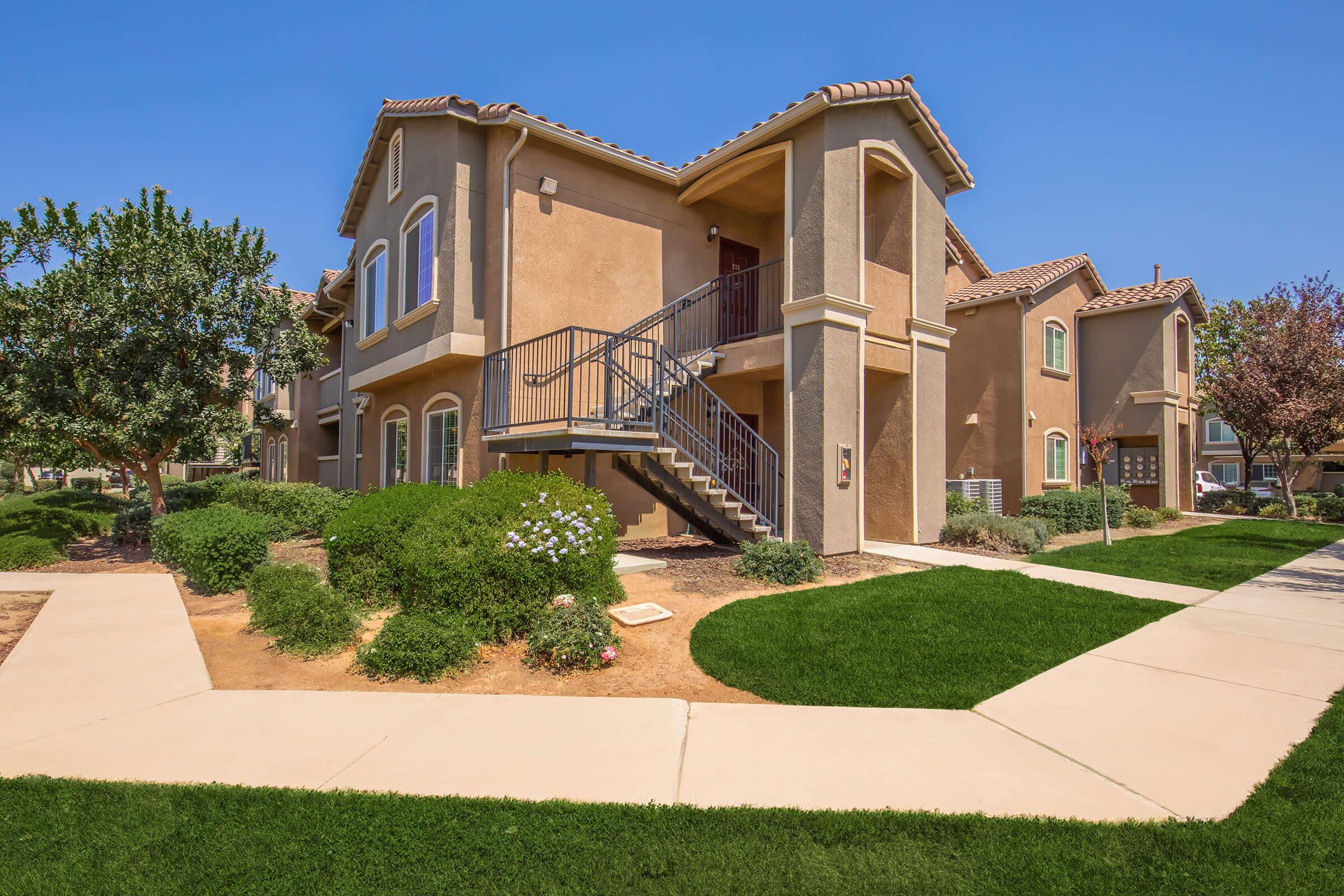 A two-story apartment building with a staircase on the side is surrounded by well-maintained greenery and landscaped pathways. The sunny blue sky enhances the inviting atmosphere of the residential area.