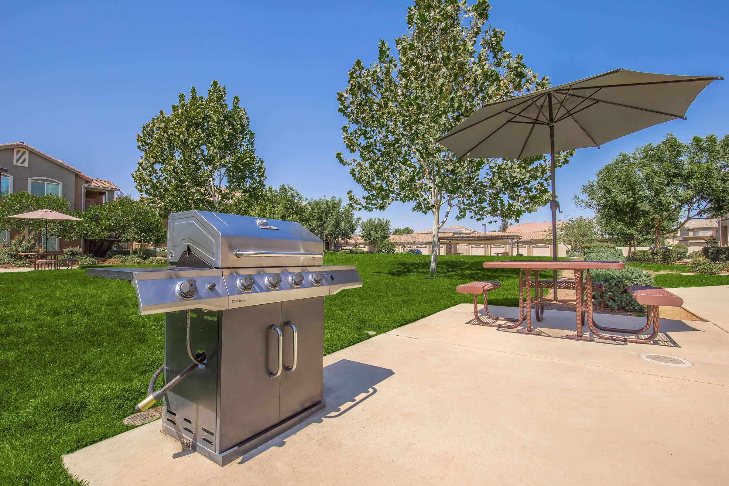 A stainless steel gas grill placed on a paved patio in a spacious backyard. Lush green grass surrounds the area, with several trees providing shade. A picnic table with benches and a large umbrella are visible in the background, creating a welcoming outdoor dining space. Clear blue skies enhance the inviting atmosphere.