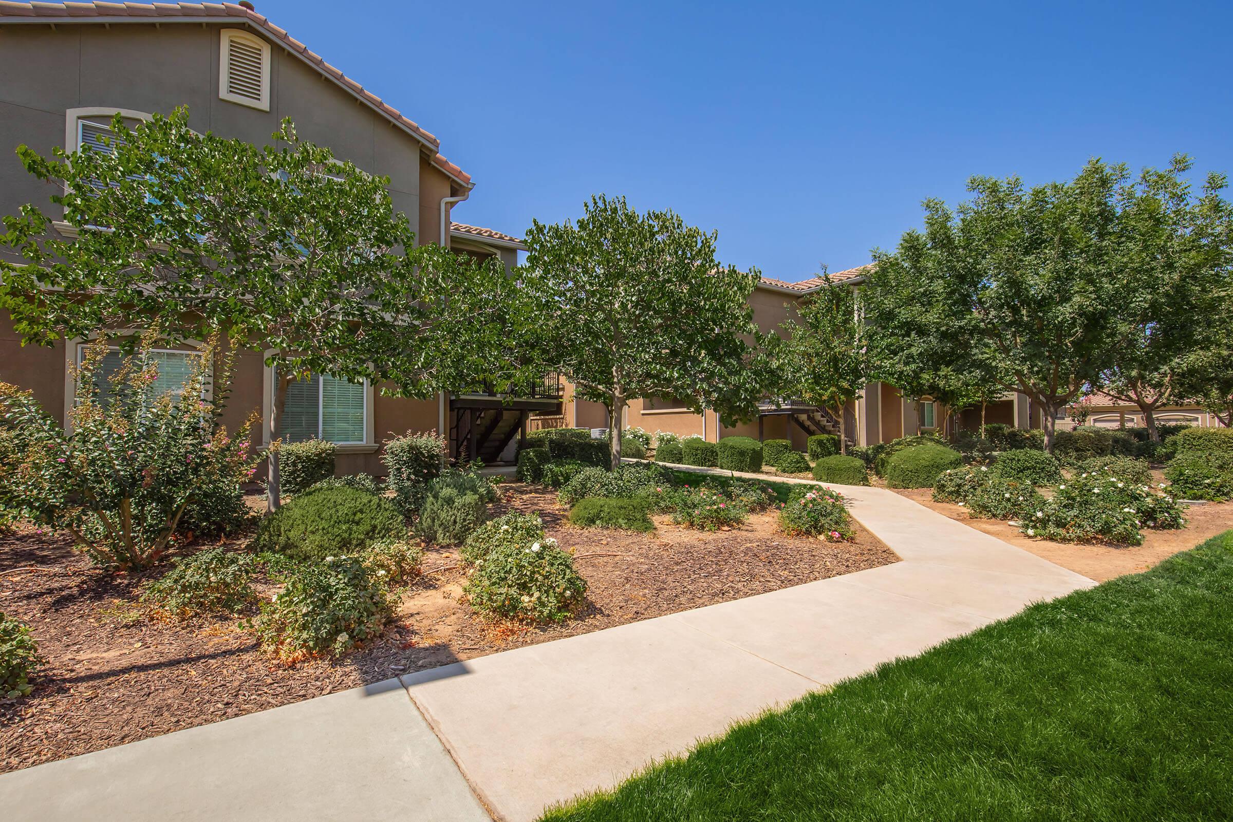 A landscaped pathway leading through a garden area with shrubs and flowering plants, flanked by residential buildings under a clear blue sky. The scene features green grass, well-maintained bushes, and a neat sidewalk, creating a serene outdoor environment.