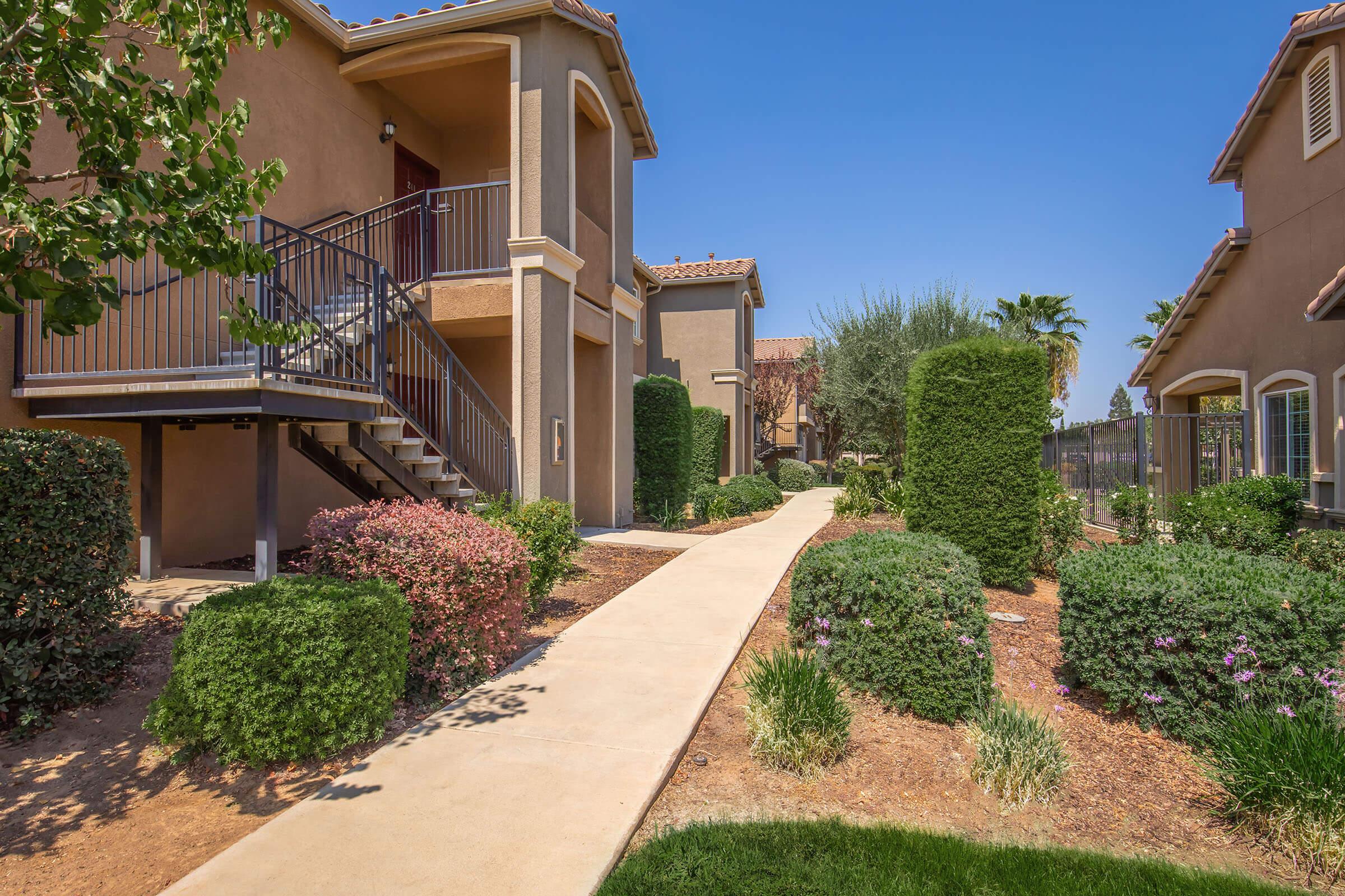 A well-maintained pathway leads through landscaped gardens with shrubs and flowering plants, lined by two-story residential buildings. The blue sky above and the lush greenery create a serene atmosphere, ideal for outdoor living. The scene reflects a vibrant, welcoming community setting.