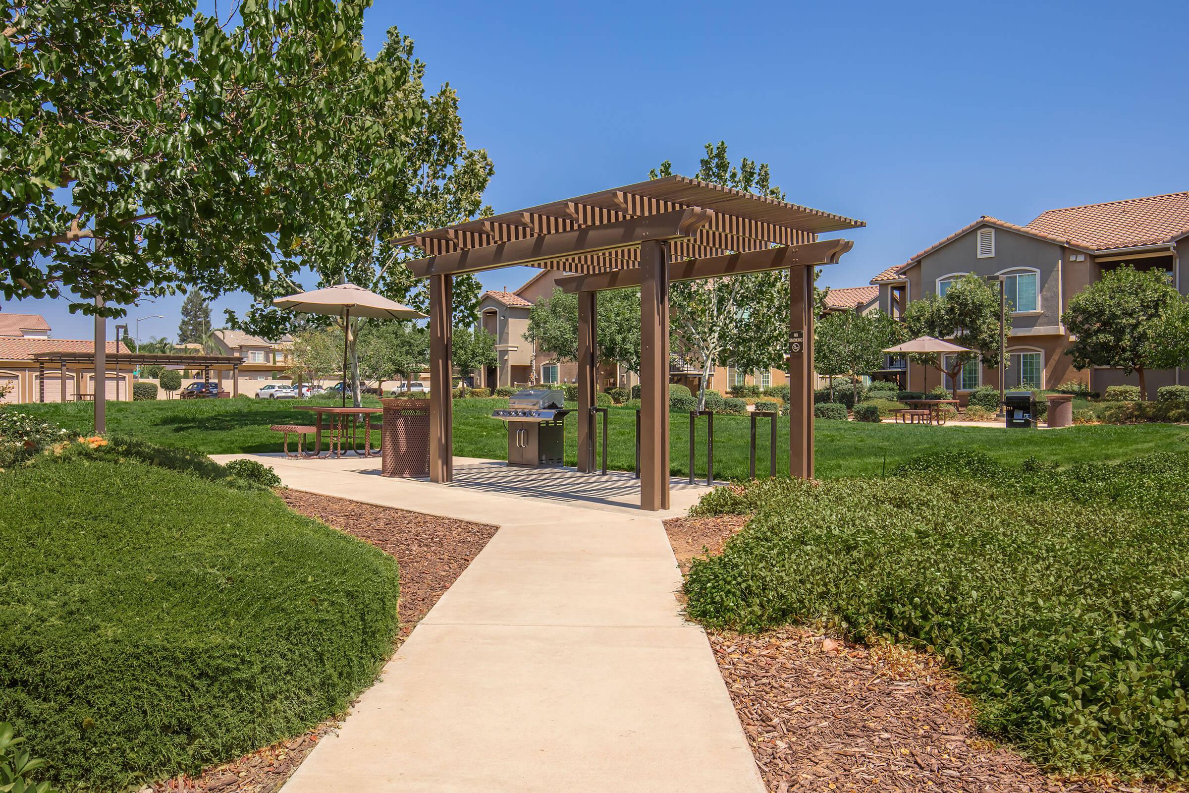A landscaped outdoor area featuring a wooden pergola with a barbecue grill underneath. The path leads through well-maintained greenery, with trees and shrubs surrounding the space. Nearby, residential buildings are visible under a clear blue sky, creating a welcoming community atmosphere.