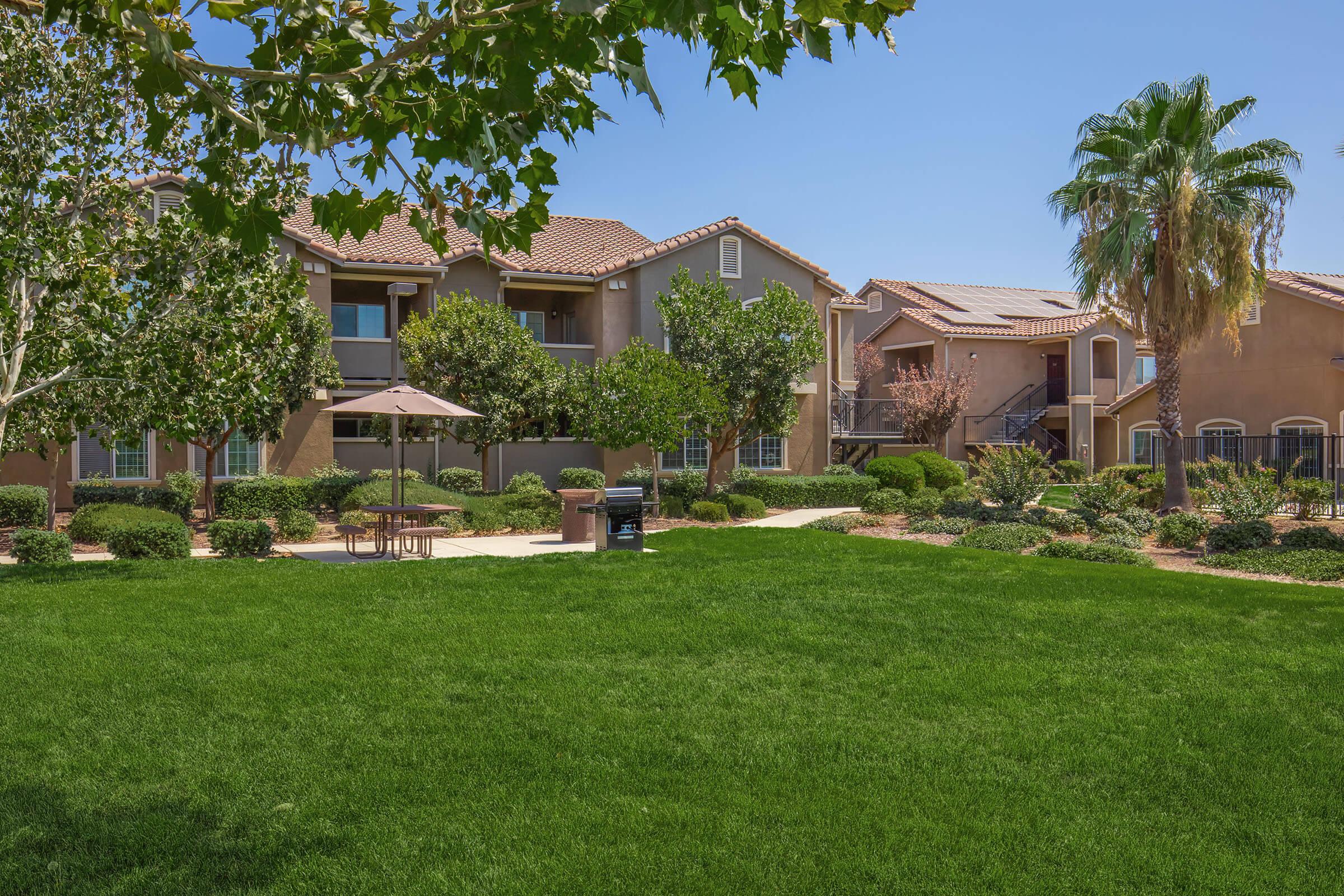 Lush green lawn in the foreground with landscaped gardens. In the background, two multi-story buildings with brown stucco exteriors and tiled roofs. There are several trees providing shade and a patio area with a table and umbrella. Clear blue sky overhead.
