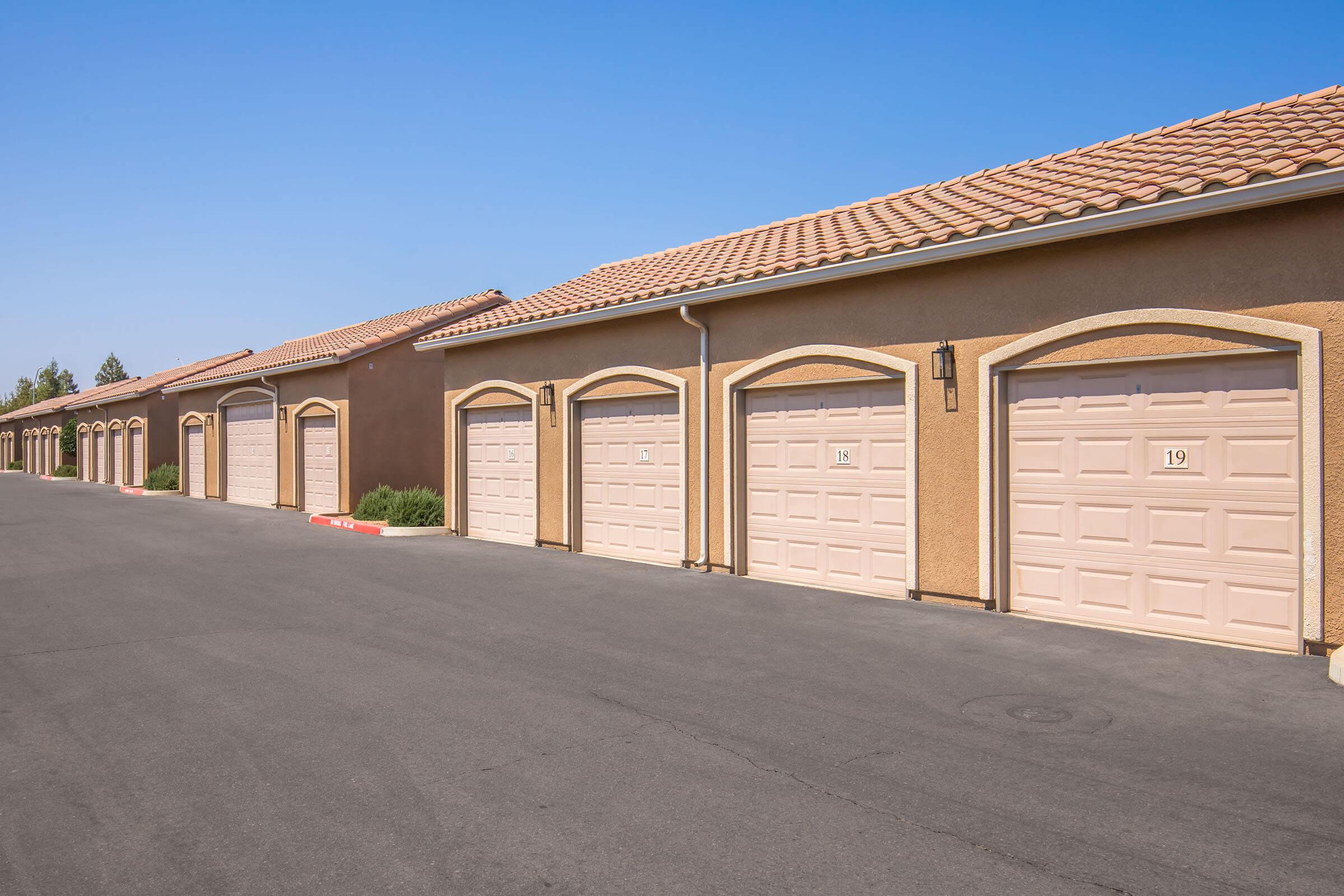 A row of beige garage doors lined up along a paved driveway, with a clear blue sky overhead and a few shrubs in the background. Each garage features an arched design and is numbered, creating a uniform appearance in a residential area.