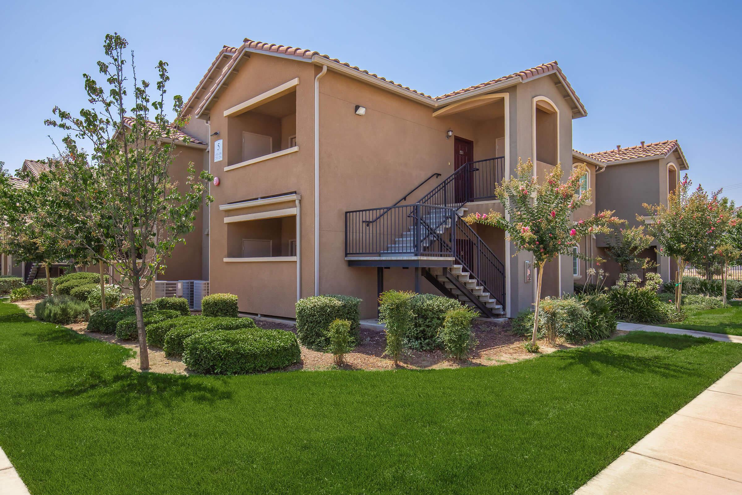 Two-story apartment building with a staircase, surrounded by well-maintained green landscaping and evenly trimmed hedges. A clear blue sky is visible above, enhancing the inviting atmosphere of the neighborhood.