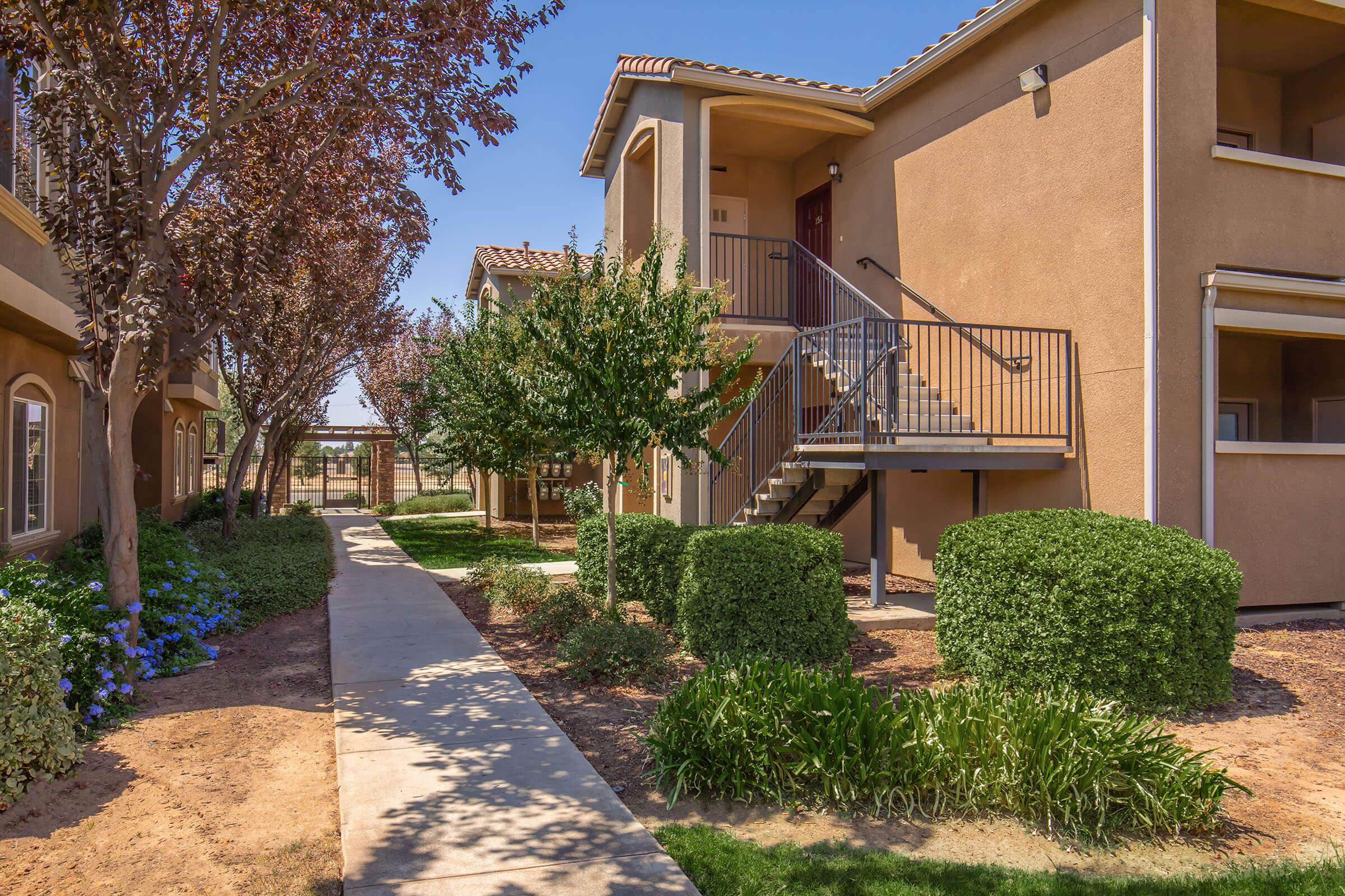 A well-maintained residential pathway flanked by manicured shrubs and flowering plants, leading to a two-story apartment building with a staircase. The scene features vibrant greenery, a clear blue sky, and a peaceful atmosphere, suggesting a pleasant living environment.